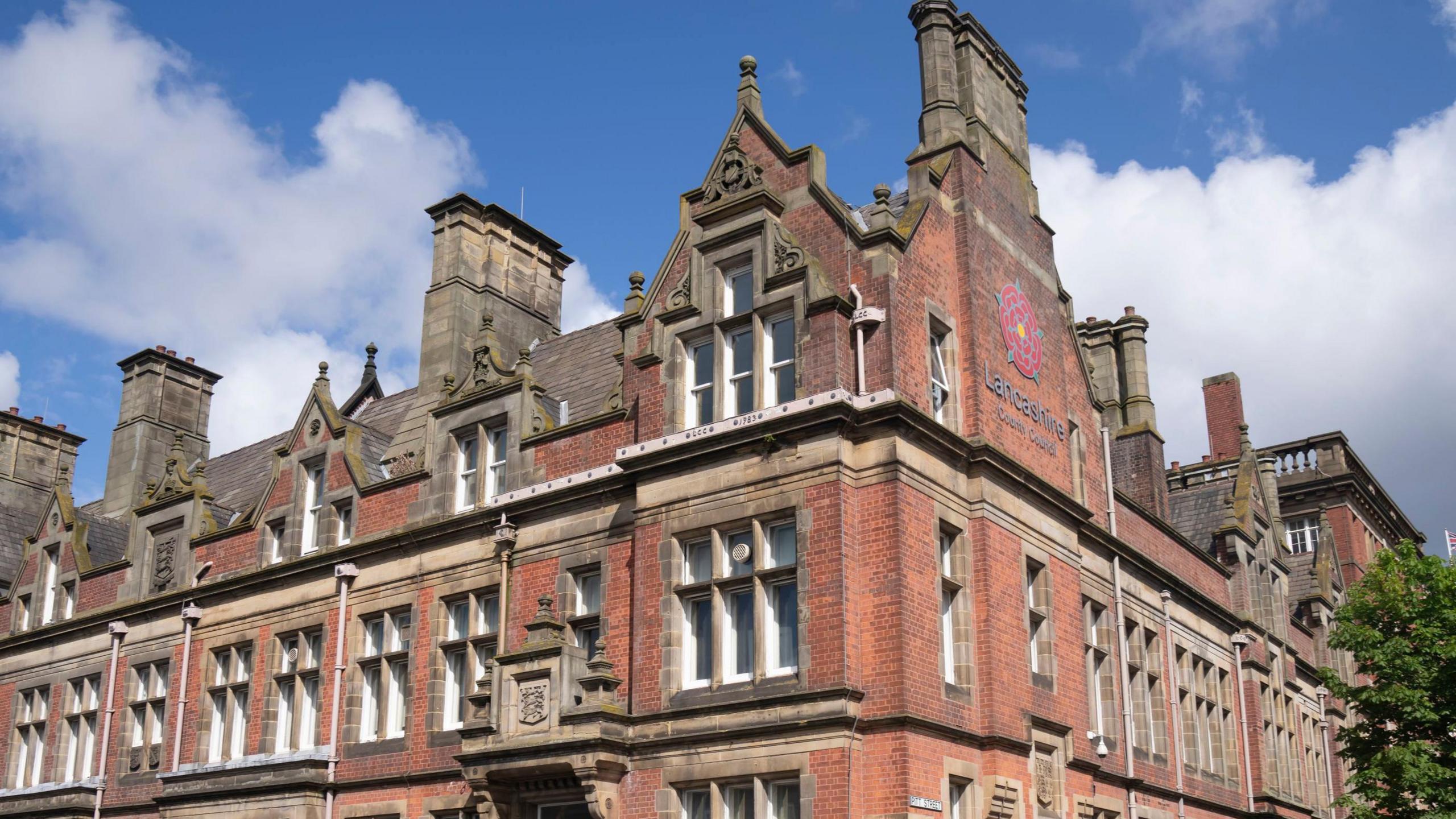 County Hall in Preston, a red-brick building with a Lancashire County Council sign on the right hand side of the building.
It's a sunny day with a few clouds in the sky.