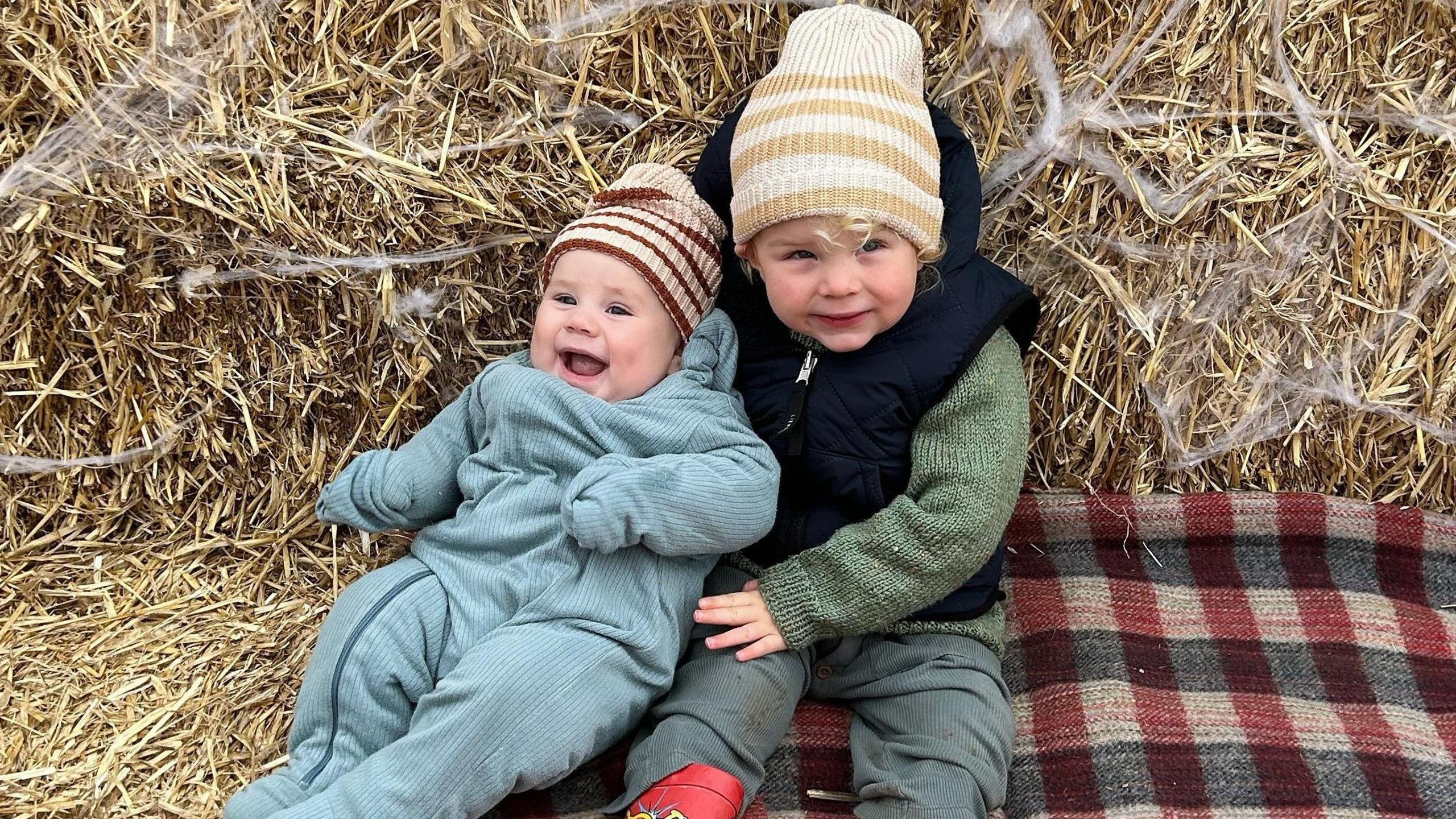 Baby being propped up by his three year old brother sitting on hay stacks and a blanket at a Halloween themed farm 