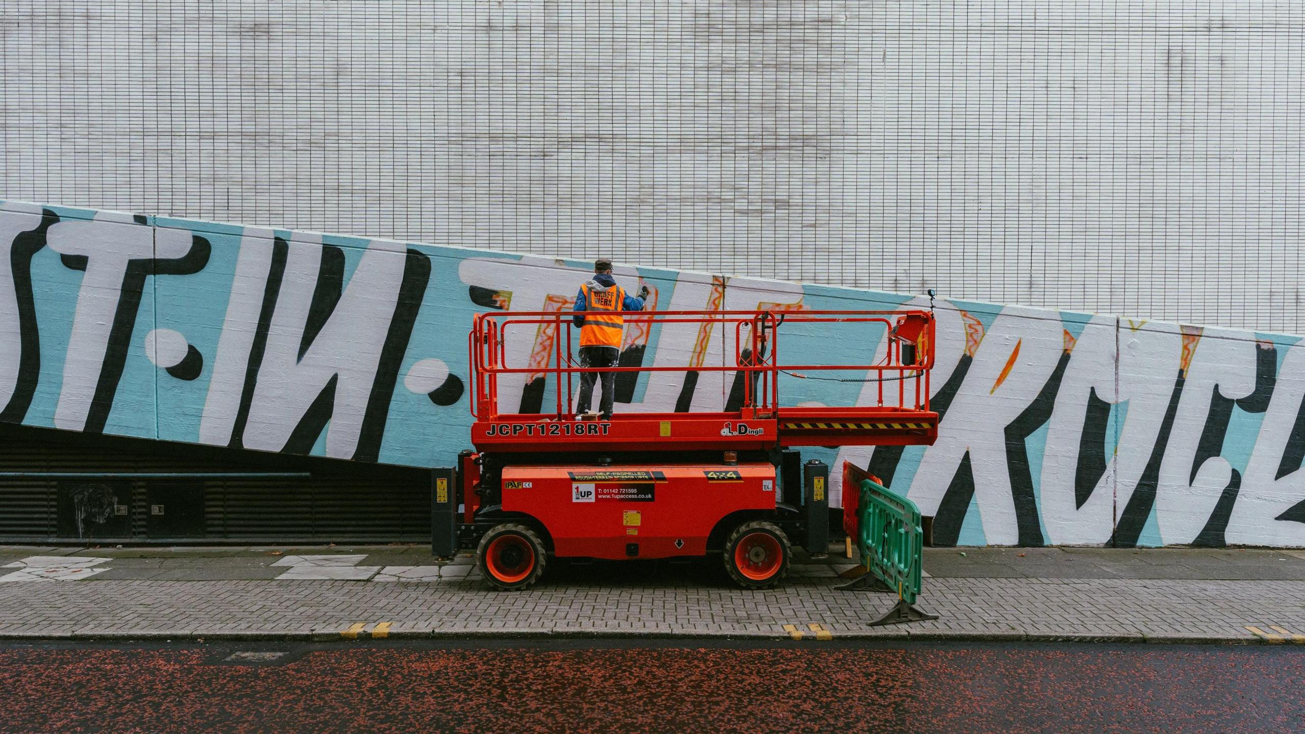 A man wearing a high-visibility orange vest stands on a red mobile elevated work platform in front of a large tiled wall. The wall features a bold mural with large black and white letters on a light blue background. The platform is positioned on a pavement next to a green safety barrier.
