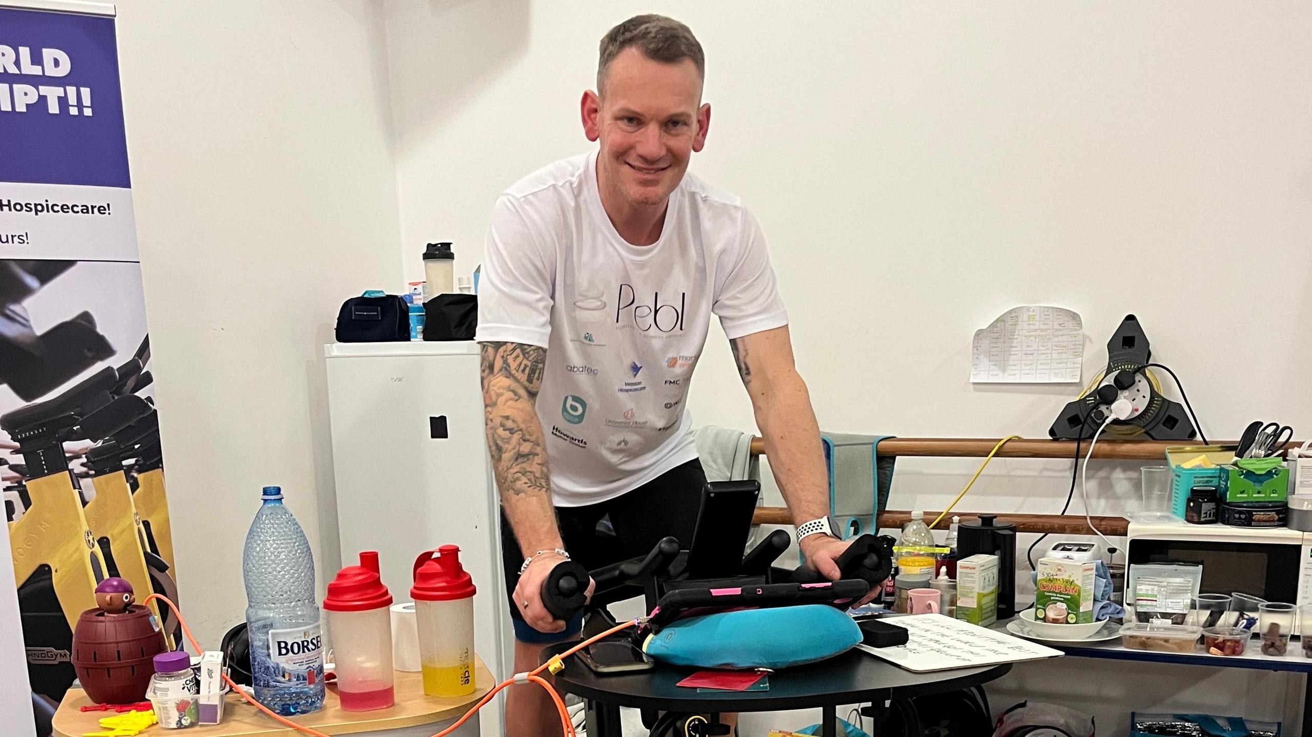 Paul on a static bike wearing a white t-shirt with short dark hair smiling at the camera