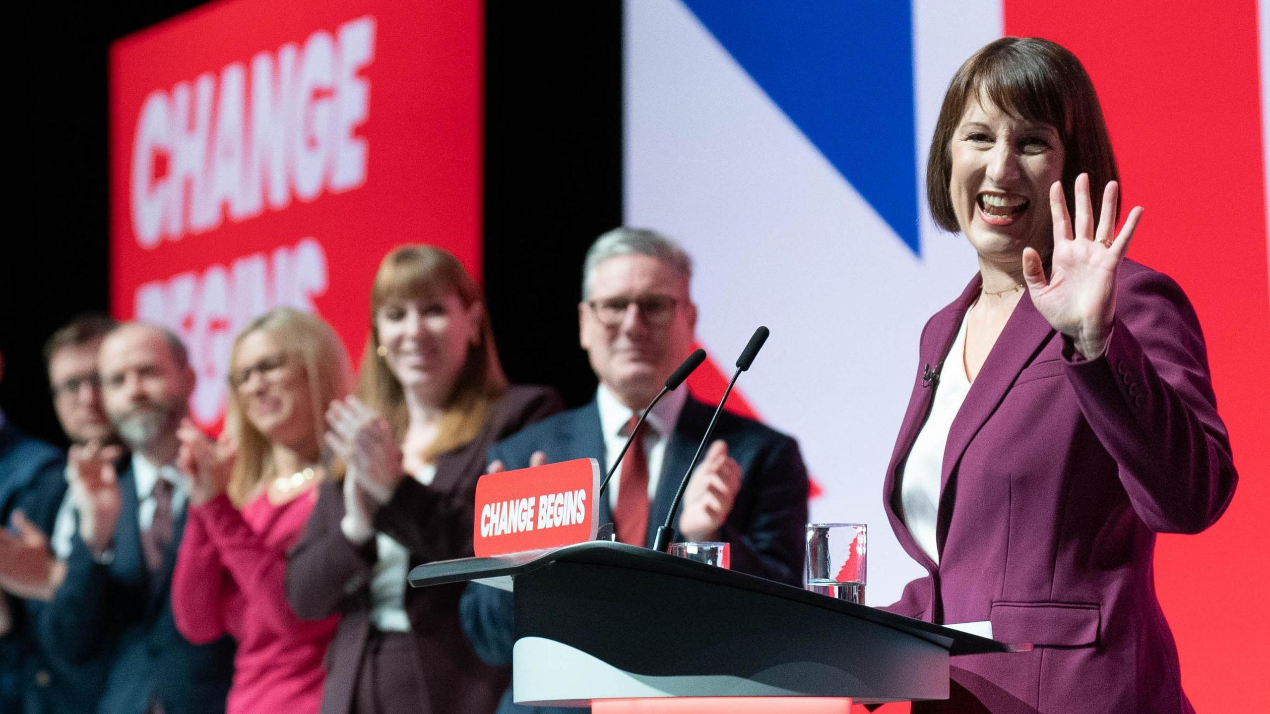 Rachel Reeves smiling and waving on stage at Labour conference, standing alongside a row of other government members including Keir Starmer and Angela Raynor against a Union Jack backdrop