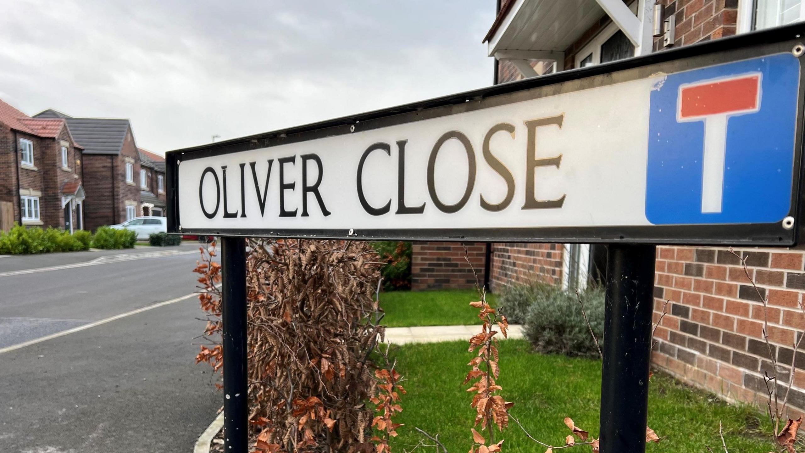 White street sign with the words Oliver Close written in black. It has a red and white 'T' sign indicating a dead end. There are houses and cars in the background.