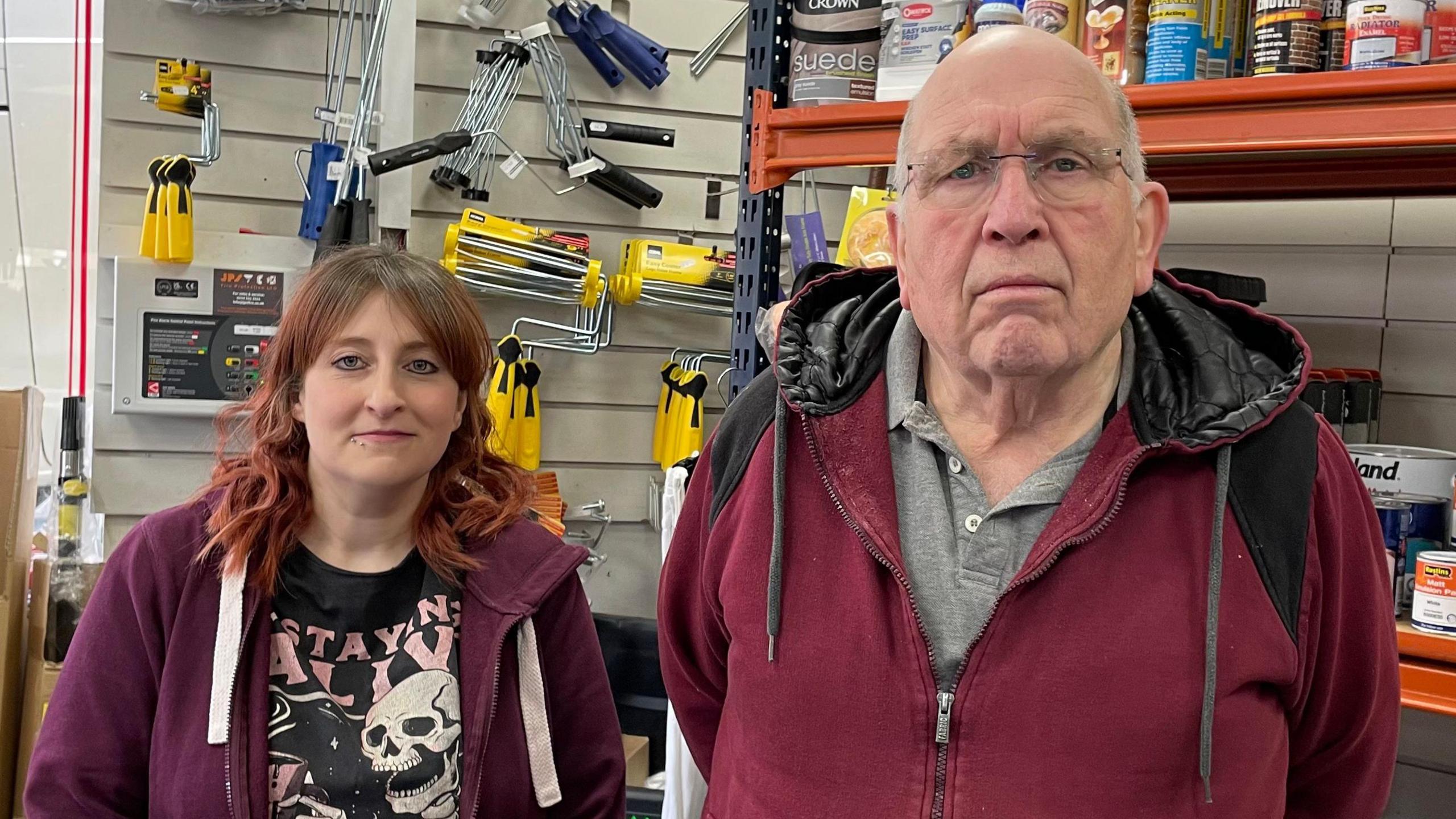 Lauren and Tony, frowning, inside the Yorkshire Decorators Centre shop, with paint tins and rollers in the background.