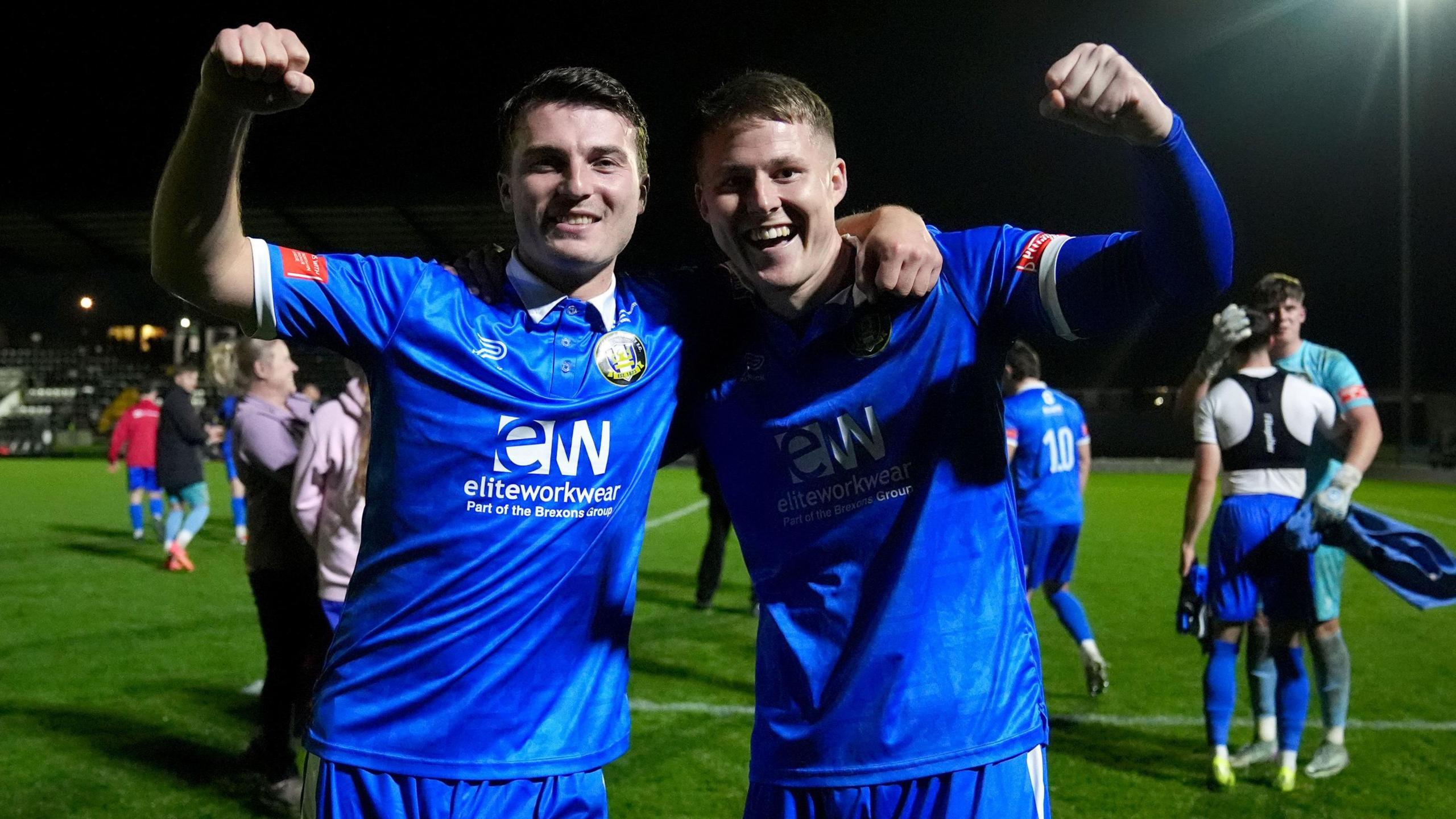 Two footballers celebrate with arms raised on a football pitch after a victory in the FA Cup. They are wearing blue shirts and shorts with a white sponsor's logo.