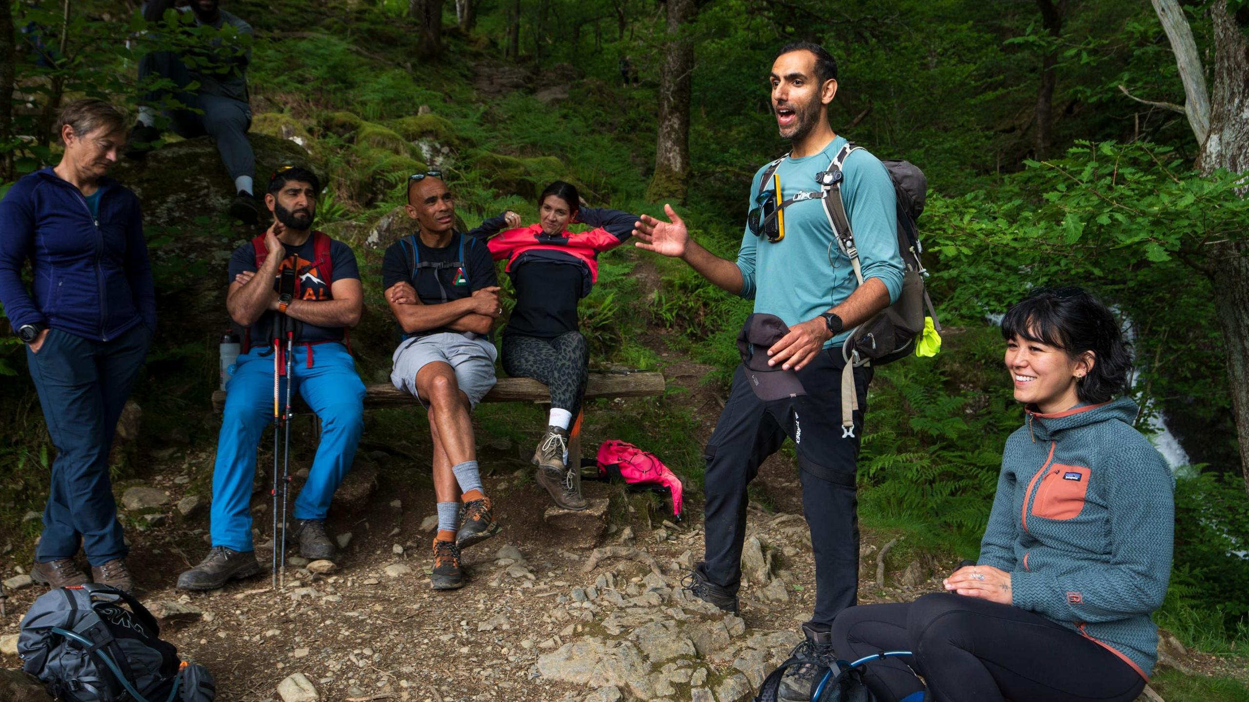 Mr Asghar is standing in a green wooded area, talking to a group of people. There is one woman standing up, two men and a woman sitting on a wooden bench, and a woman sitting on the ground smiling.