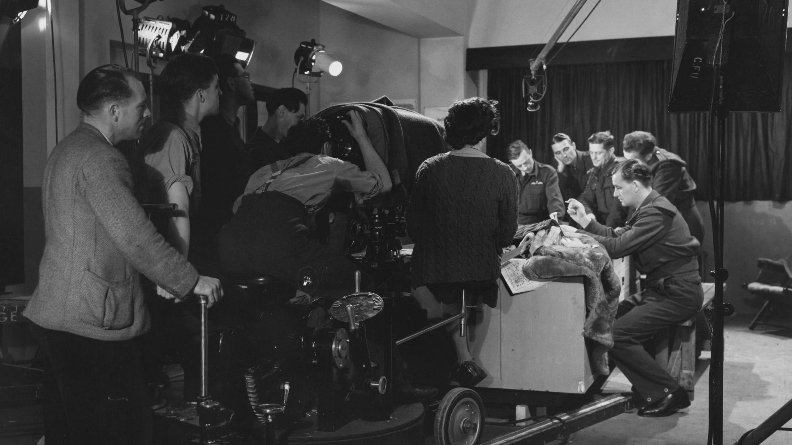 A crew is gathering round a big camera, with their backs to the person taking this photo. In front of them, are men in military uniform gathering round a table. They appear to be in a film studio.