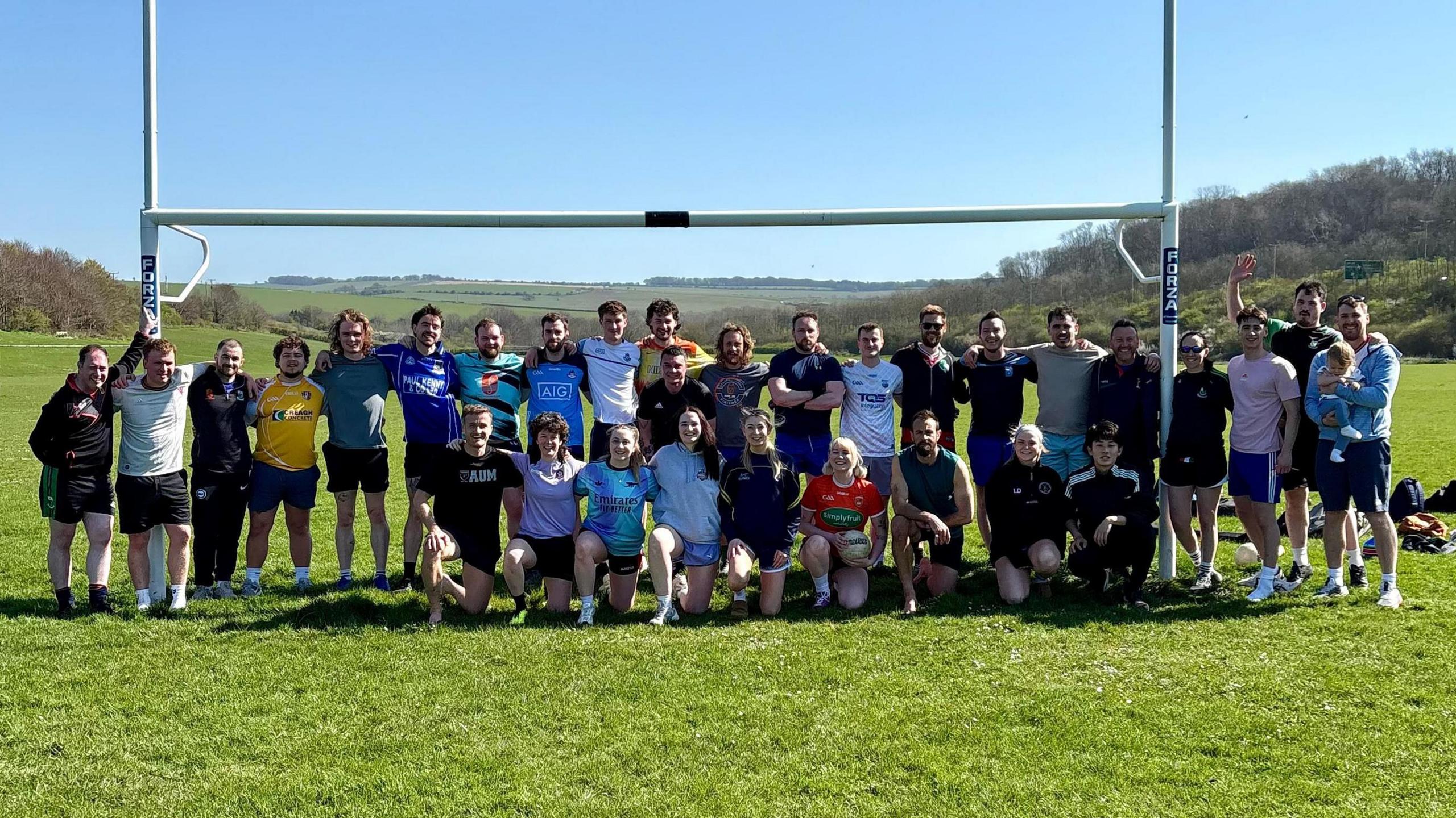 Members of Brighton & Crawley Gaels' Gaelic football team pose at their Waterhall training site in Brighton. They are standing in front of a white Gaelic football goal, featuring a goal frame with two taller posts towering either side of the crossbar. It is a clear blue day with green, rolling hills spanning the horizon.