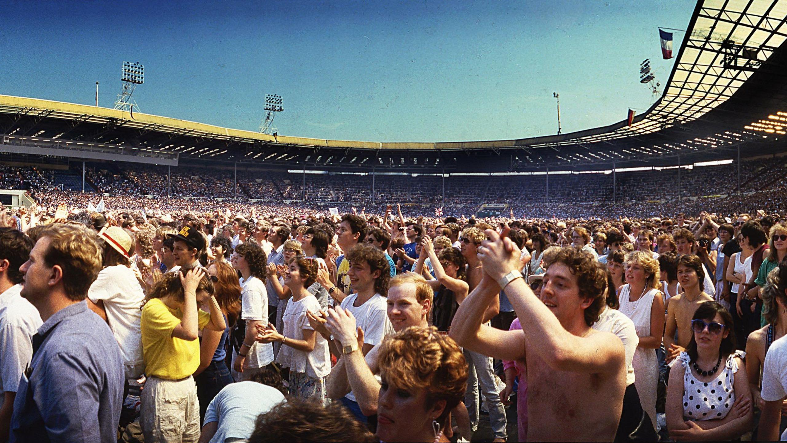A wide shot of the Wembley Stadium crowd cheering and applauding under a clear blue sky.