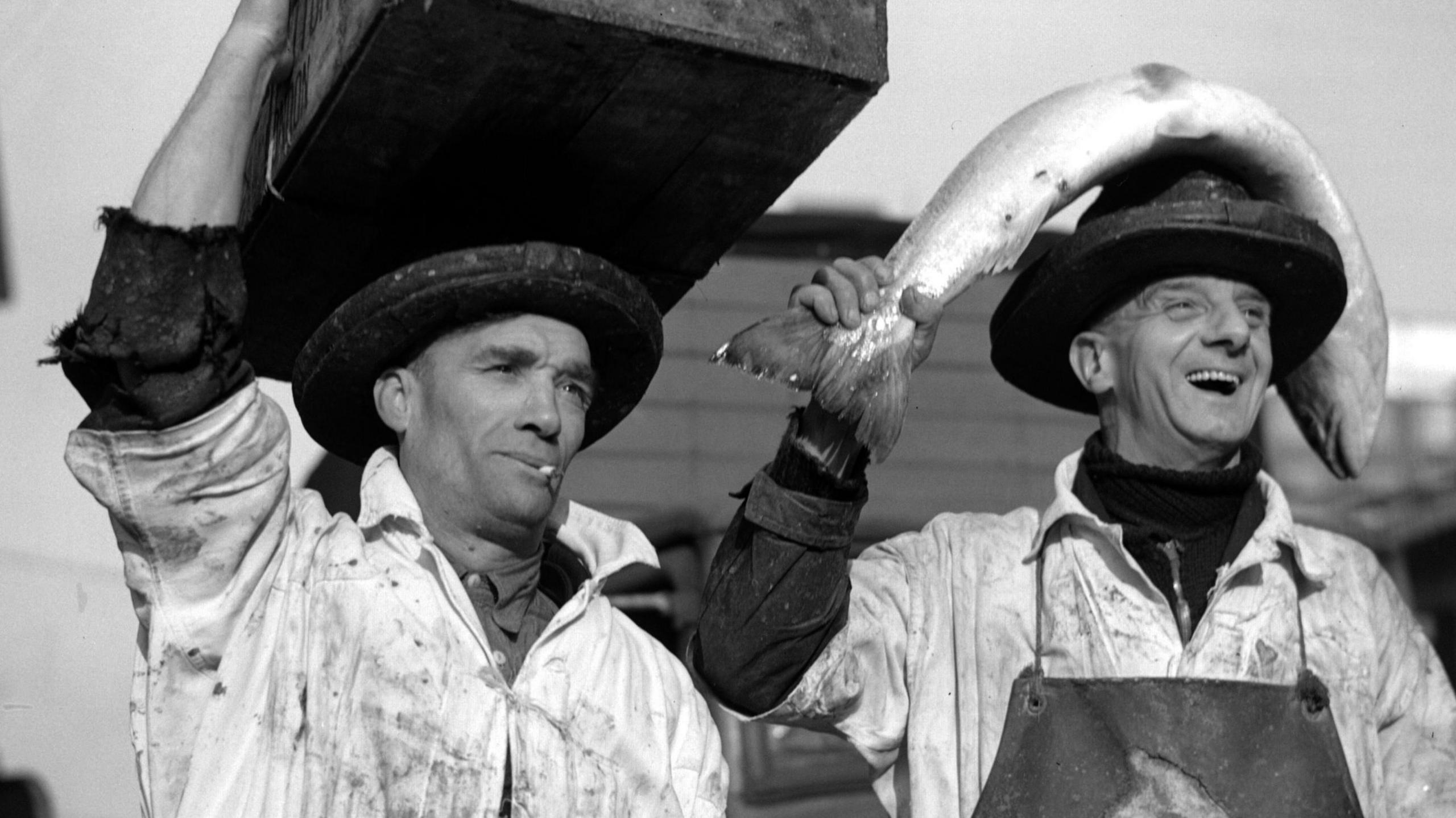 Black and white photo of two fish porters, one with a case on his head, the other with a large salmon on his head