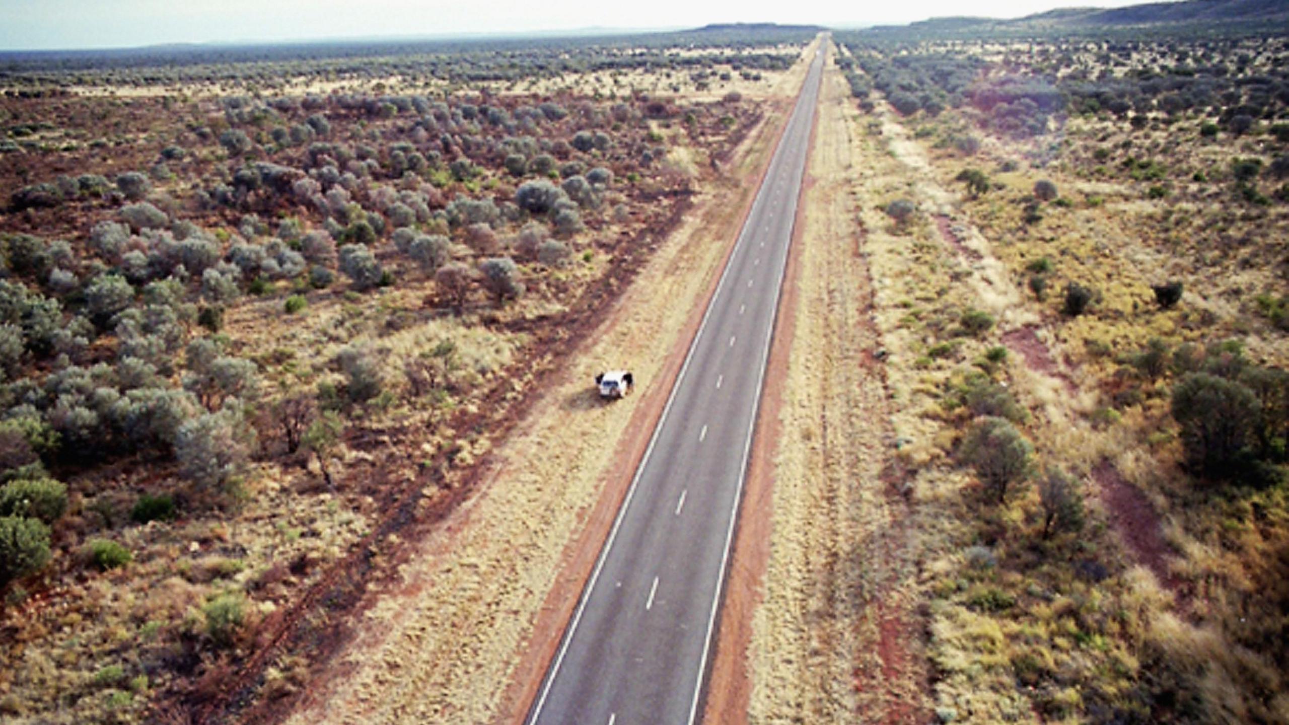 A long, straight and empty road, pictured from above. Shrubs, trees and bushland are on both sides.