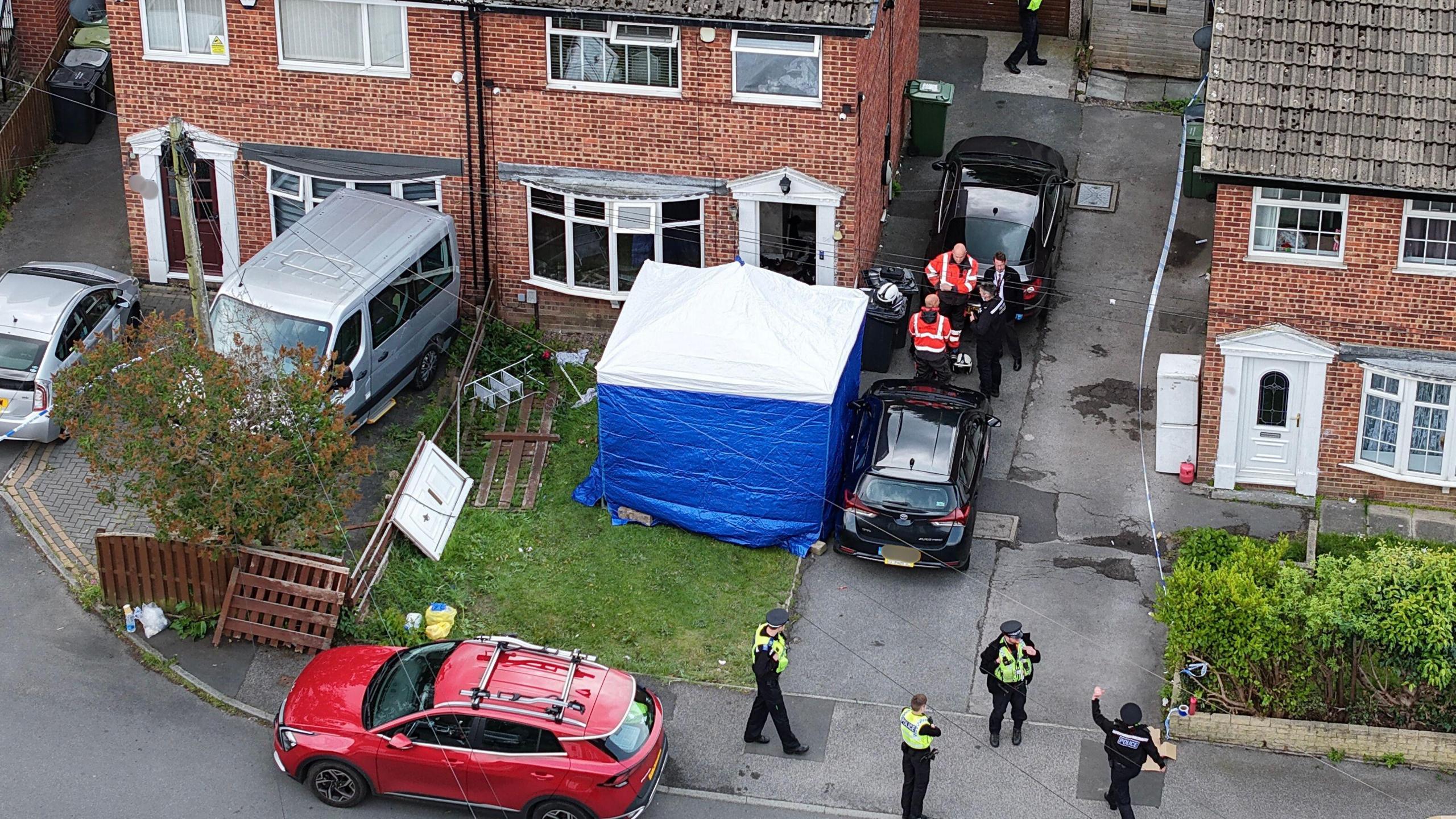 Police gather in front of a row of semi-detached properties. One house has a blue police tent in the front garden and has the door missing. 