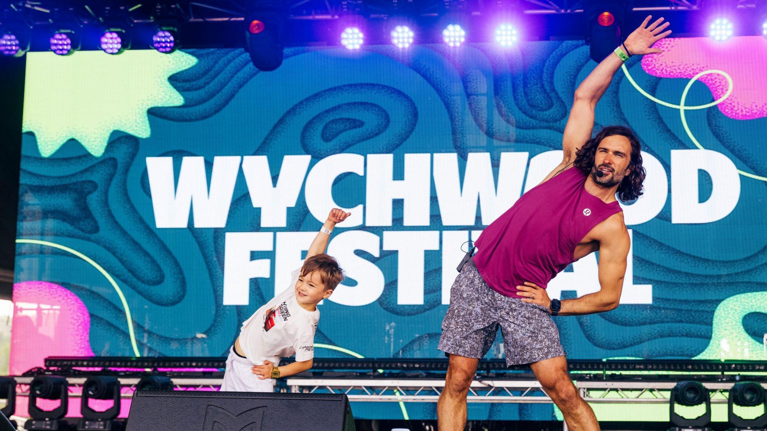 Trainer Joe Wicks performs stretches at Wychwood Festival on the main stage with a young boy from the crowd. Behind them is a large banner bearing the name of the festival