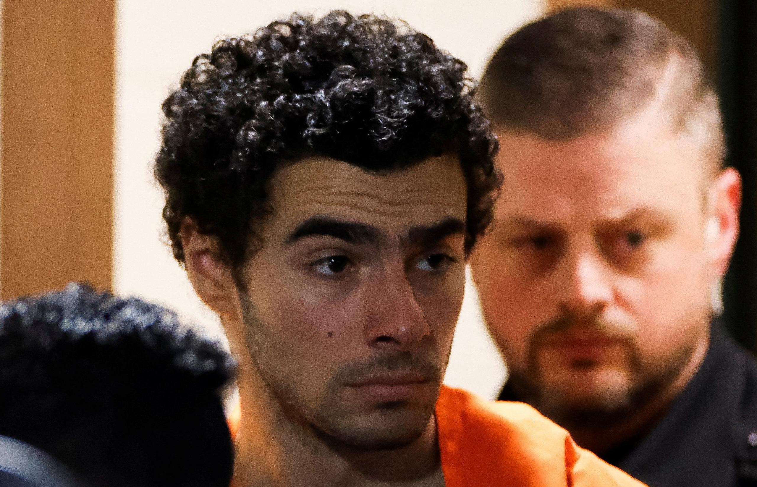 Close up photo of Luigi Mangione in an orange prison jumpsuit. Police are around him and his facial expression is serious.