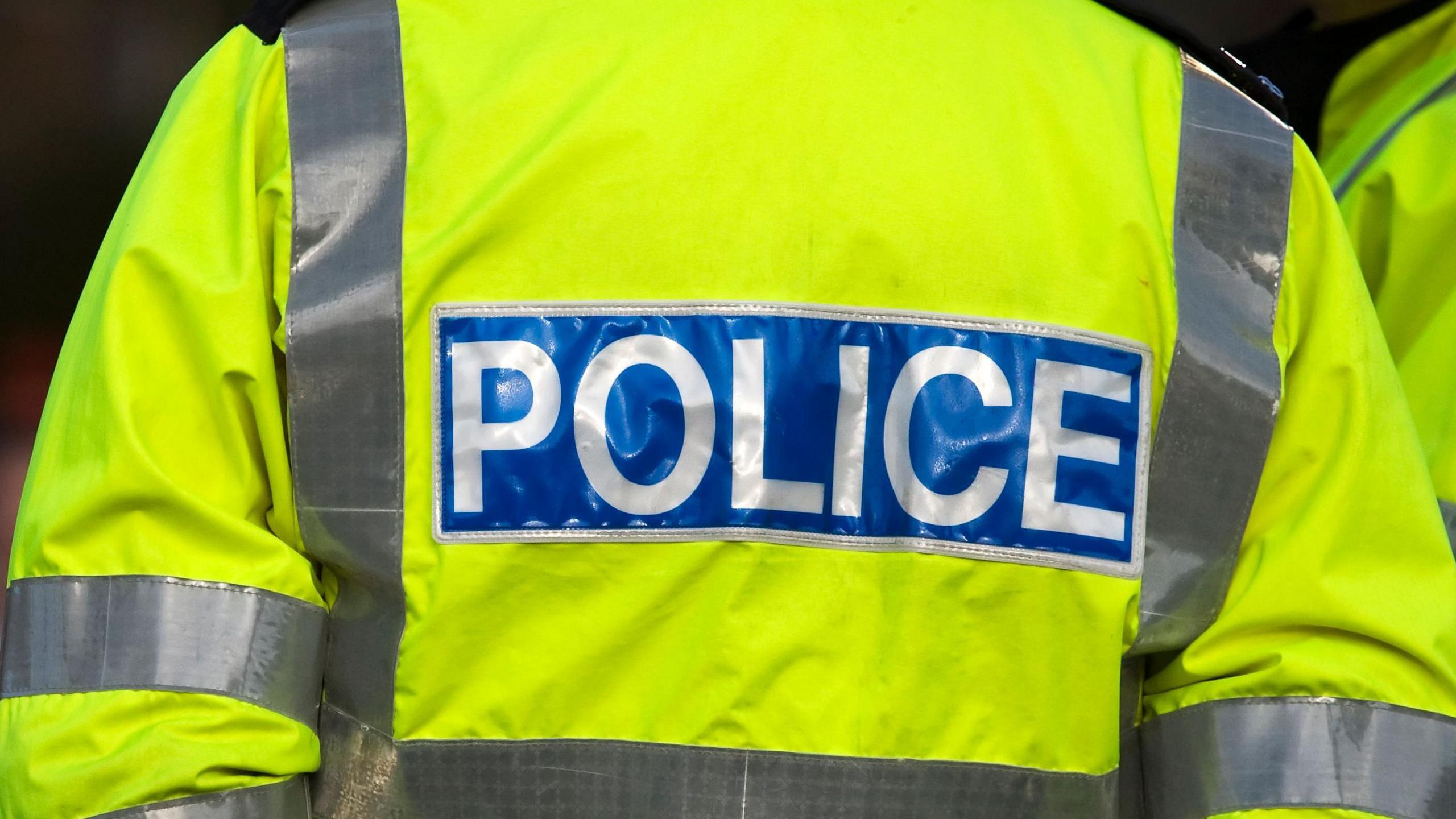 A person wearing a high-vis yellow police jacket. The picture shows them from behind. Displaying the blue police wording. 