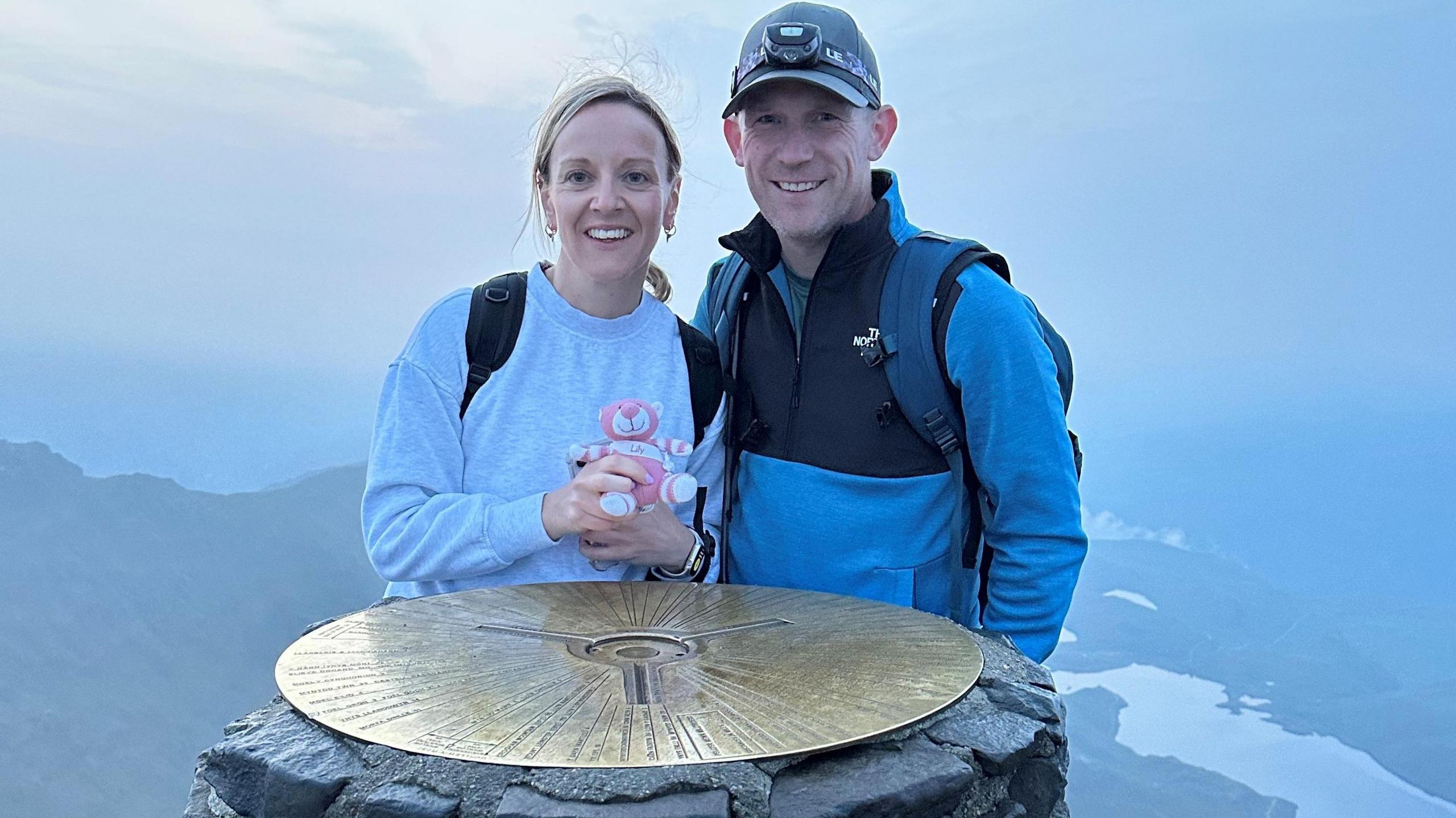 A woman and a man stand side-by-side holding a pink teddy bear. They are high up with a mountain and lake in the distance. 