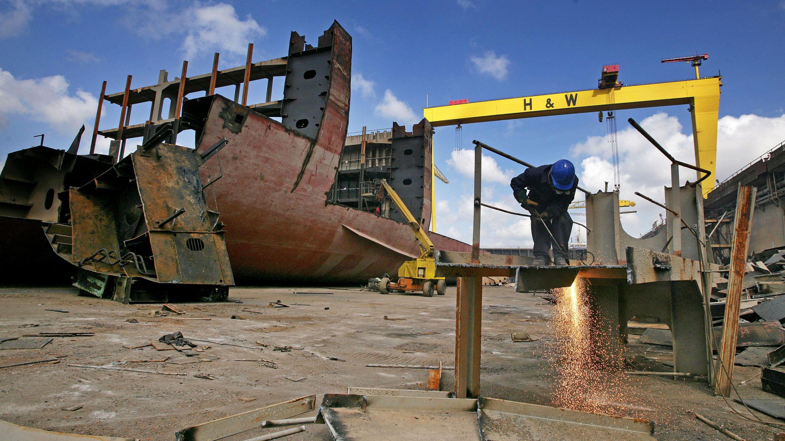 A shipyard worker, to the left of the image, cuts up pieces of the MSC Napoli cargo ship, as it lies in a dry dock at the Harland and Wolff. A yellow Harland & Wolff crane is in the background. 