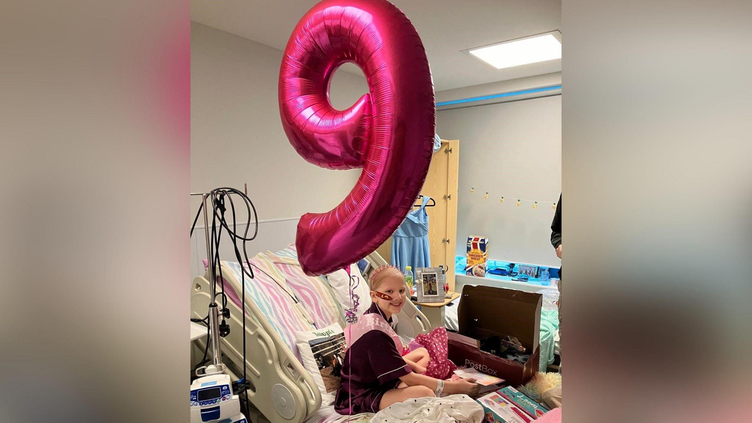 Kathryn yn gwenu wrth eistedd ar ei gwely yn yr ysbyty. Mae yna falŵn mawr binc o'r rhif naw o'i blaen hi. Mae'n gwisgo crys-t porffor.
