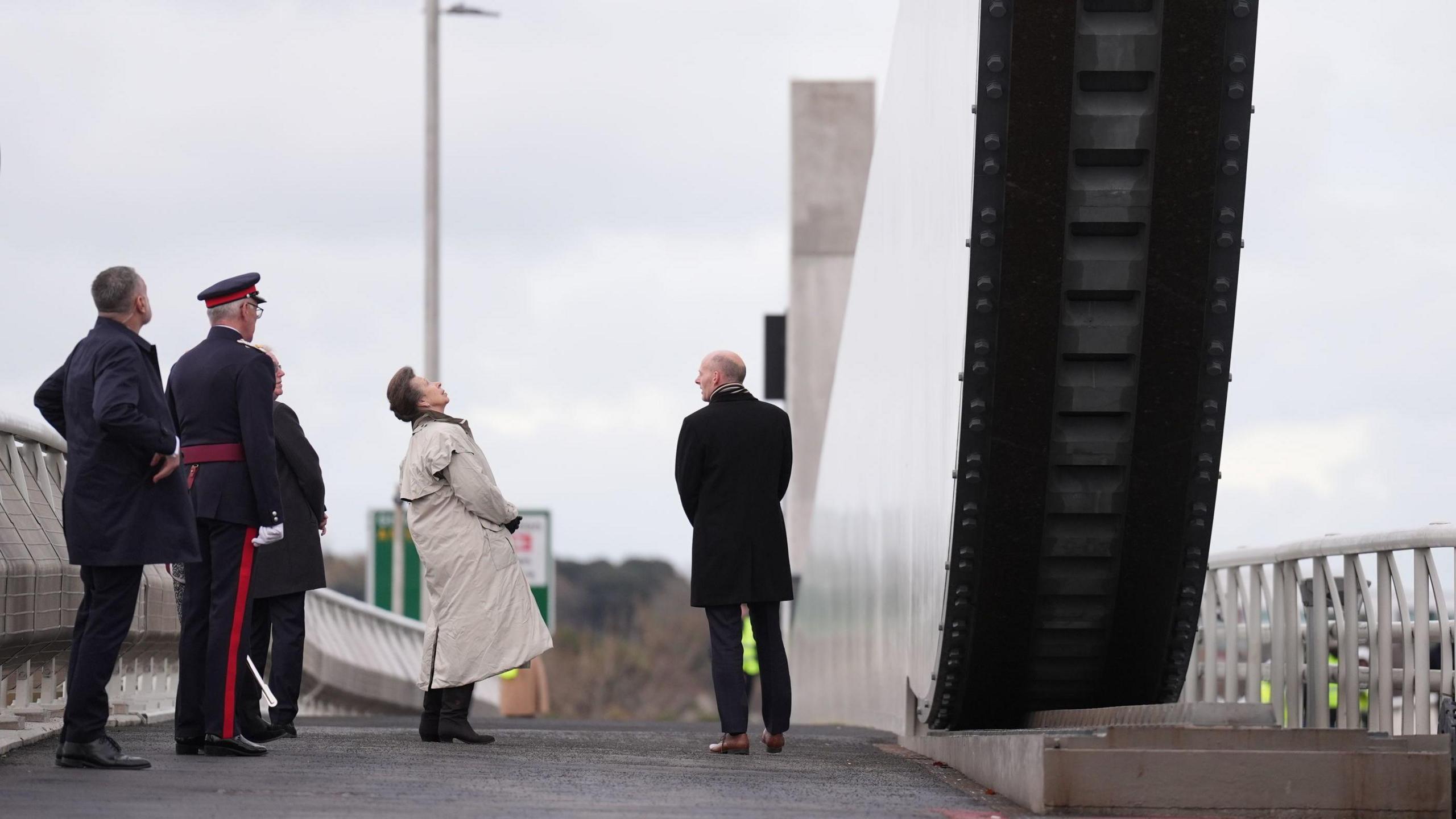 Princess Anne looks up at the bascule span on the bridge. Other members of the group that escorted her on to it stand behind her.