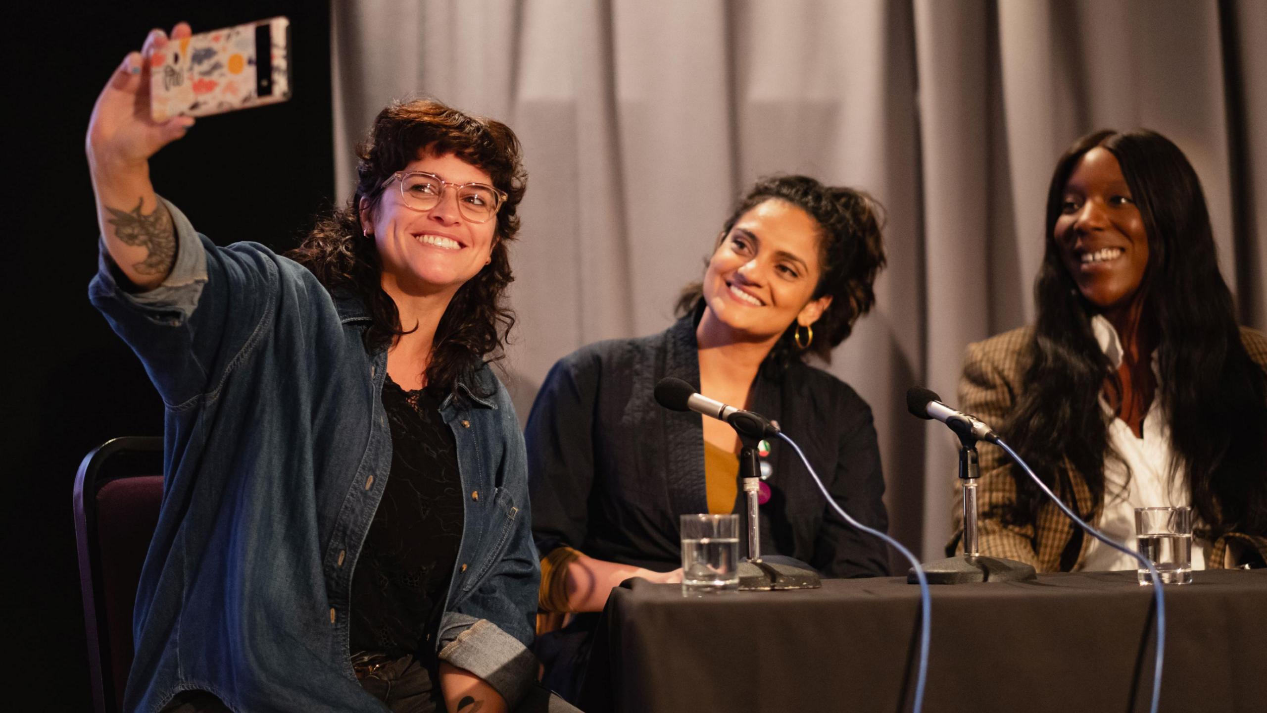 Three women sit together at a table during the Encounters Film Festival in Bristol. One of them, who has long dark curly hair and is wearing glasses, is taking a selfie to include the other two women behind her. They are both smiling and also have long dark hair.