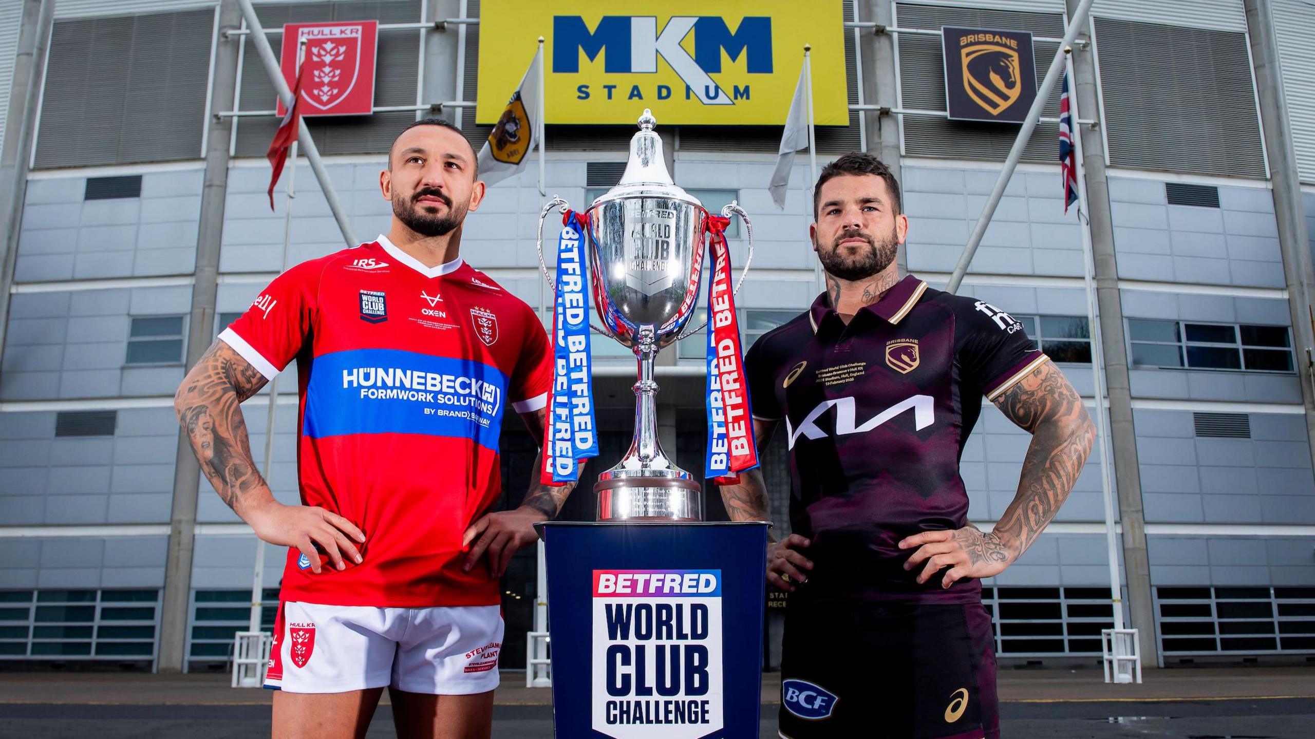 Elliot Minchella and Adam Reynolds stand either side of a plinth with the World Club Challenge trophy on it, just outside the frontage of the MKM Stadium for a promotional photograph wearing their respective Hull KR and Brisbane Broncos kits.