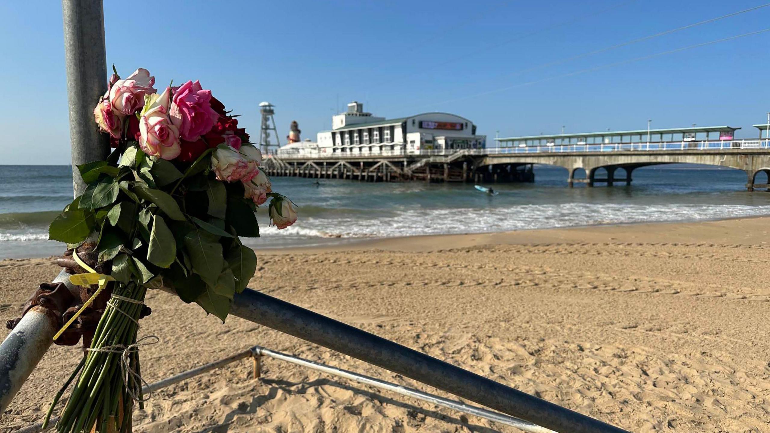 A bunch of roses is seen tied to a pole next to Bournemouth Beach, with the pier and sea appearing out of focus in the background  