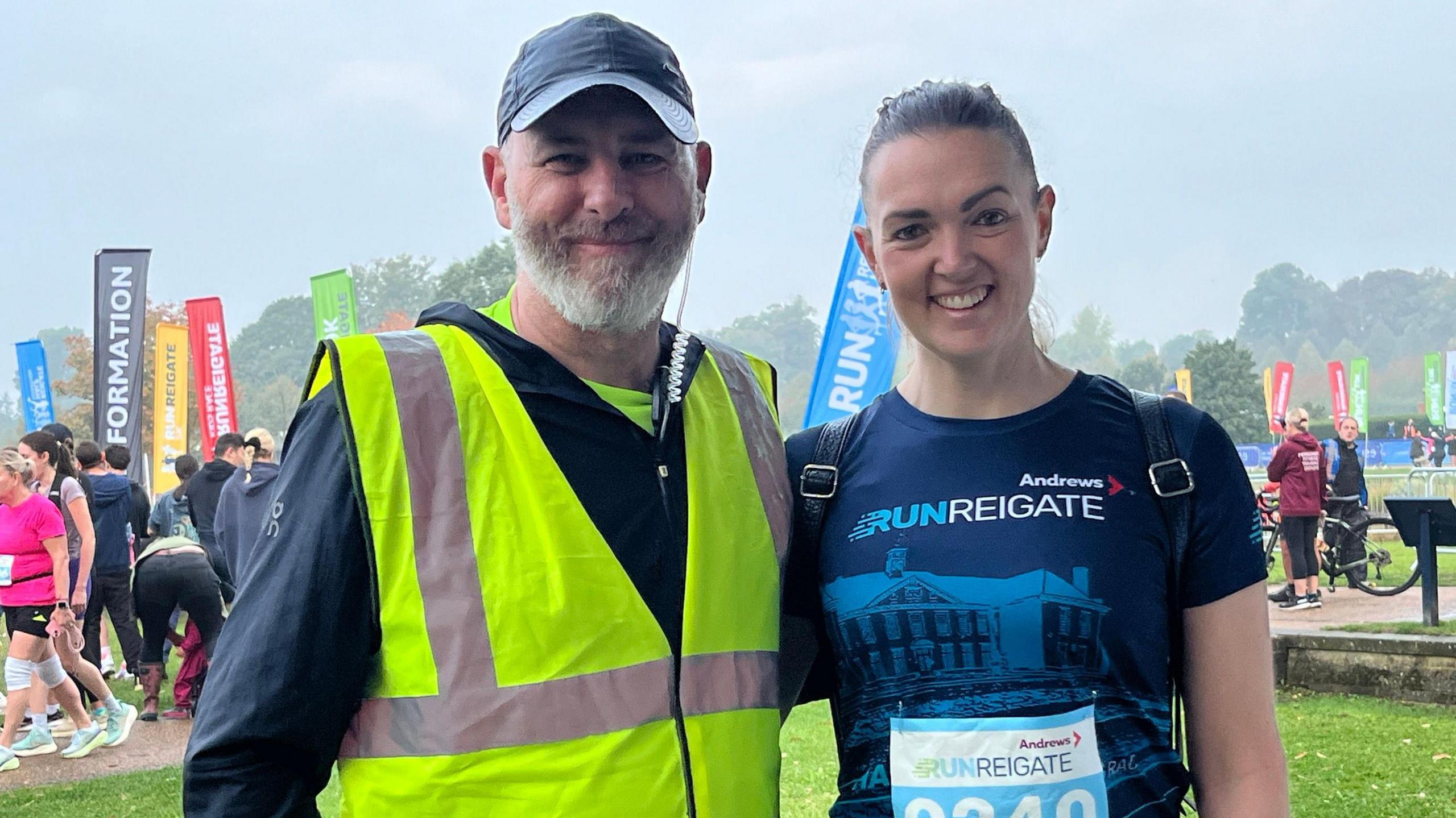 Hayley Young faces the camera, smiling, and she is wearing a dark blue t-shirt with the Run Reigate slogan across the front in white. She has dark brown hair pulled back of her face. Next to her on her right stands race director David Kelly who has a white beard and wears a dark blue baseball cap, a dark long-sleeved top and a high-vis yellow sleeveless top.