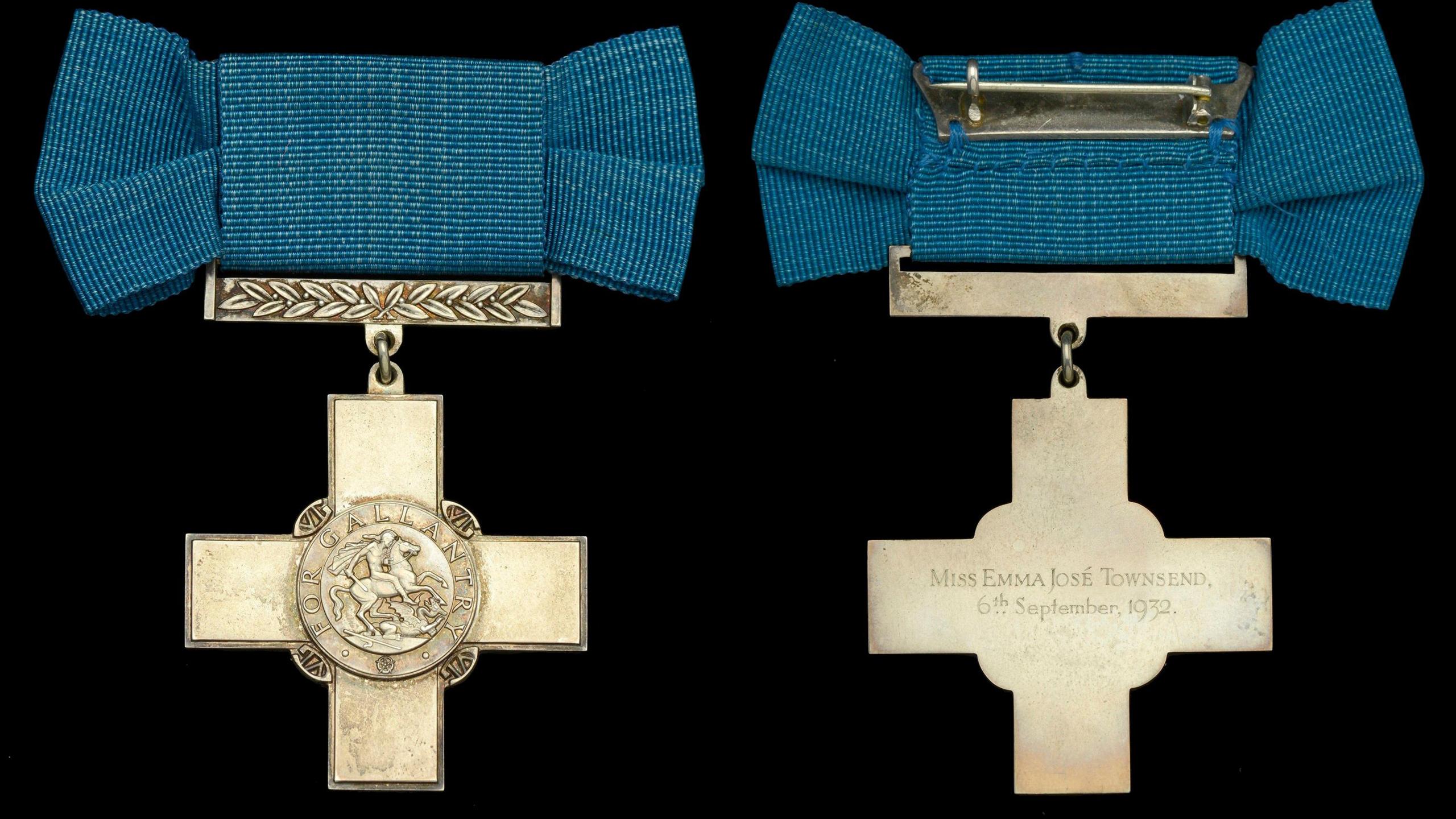 The front and back of the George Cross which is made of pale golden coloured metal mounted on a blue bow with a pin. On the front is an engraving showing a figure on a horse and on the reverse words are engraved reading 'Miss Emma José Townsend, 6th September, 1932.