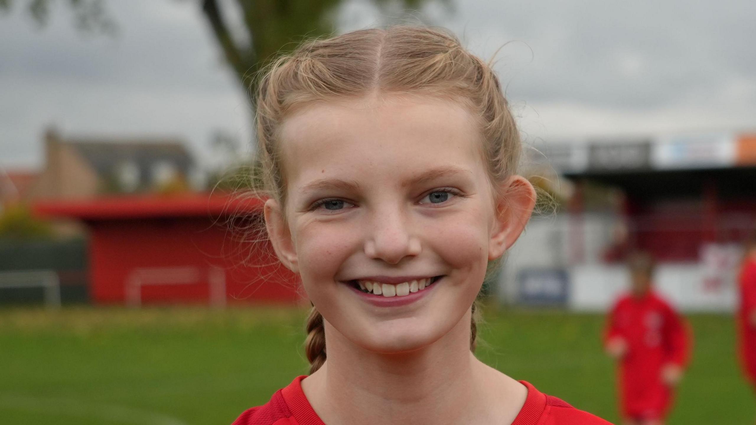 Nancy smiles at the camera while standing on a football pitch. Players can be seen behind her. She has blonde hair which has been braided on each side of her head and tied back. 