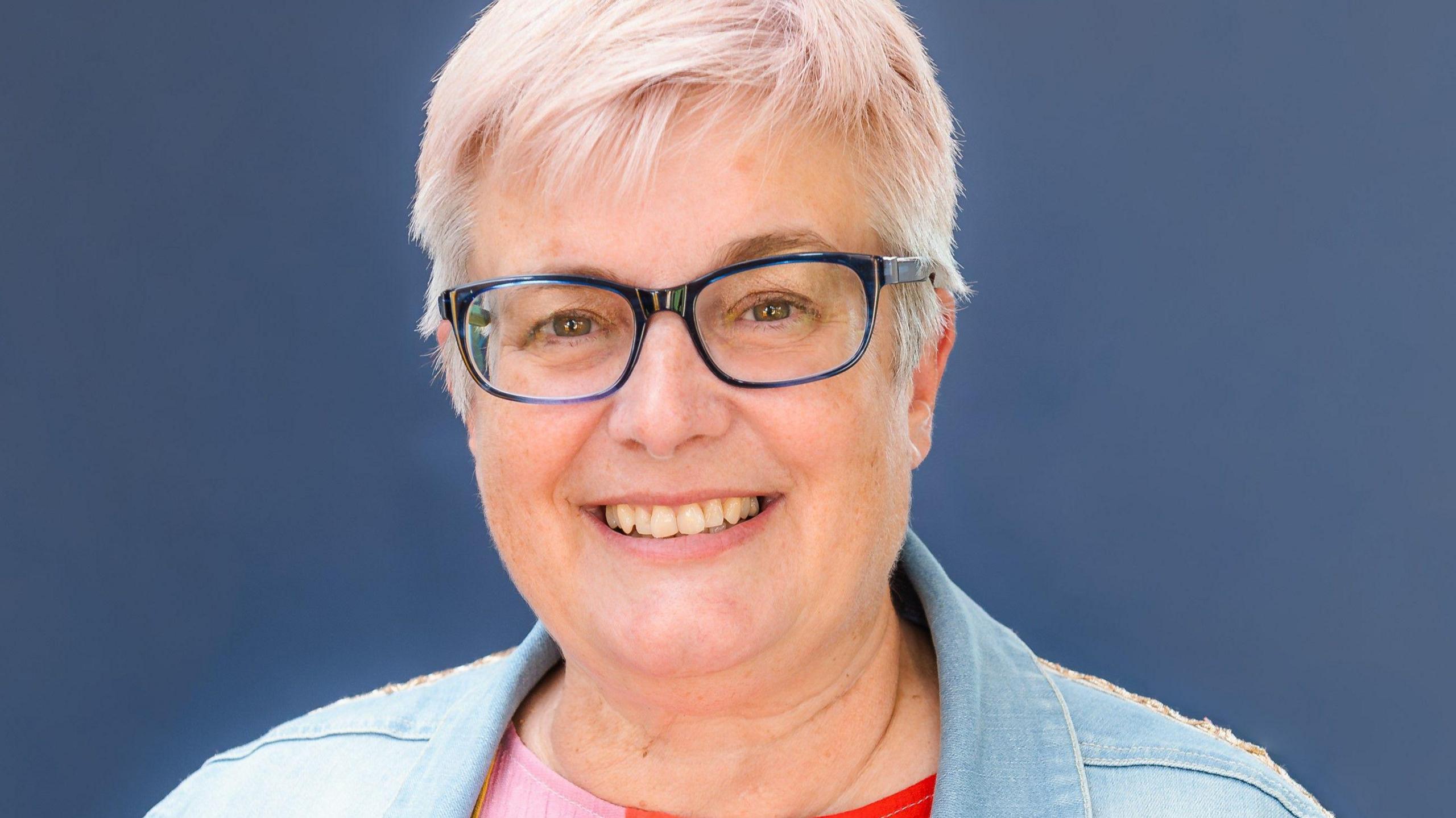 A woman with short white hair, wearing a denim jacket and glasses, is smiling at the camera. She is stood in front of a navy background