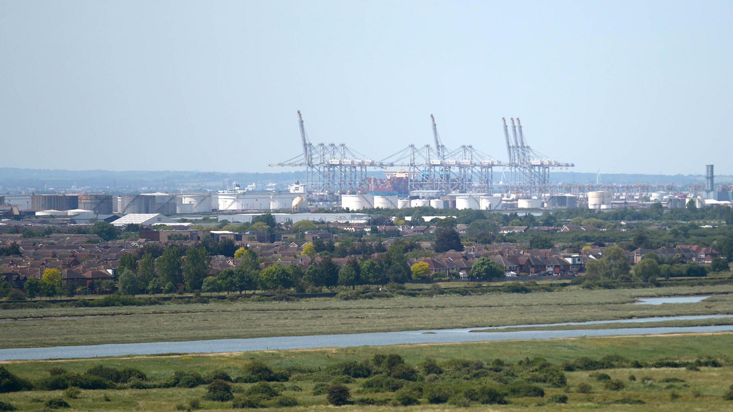 A view of the DW World Thames Gateway's cranes - visible behind the drums of a refinery, with housing, trees and marsh in the foreground