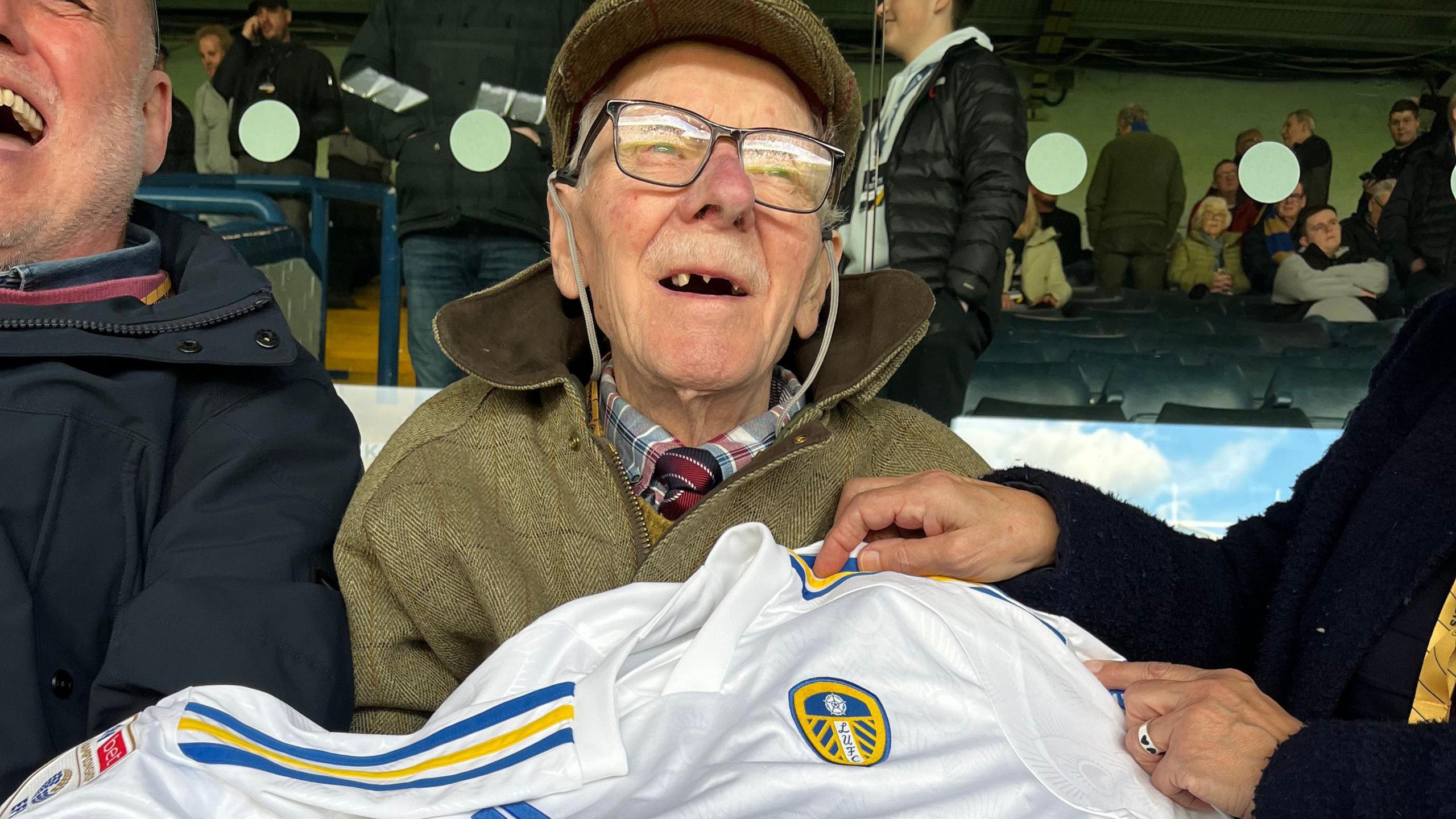 An elderly man, wearing a tweed jacket and glasses, sits in the stands of a football ground and holds a white and blue Leeds United shirt.