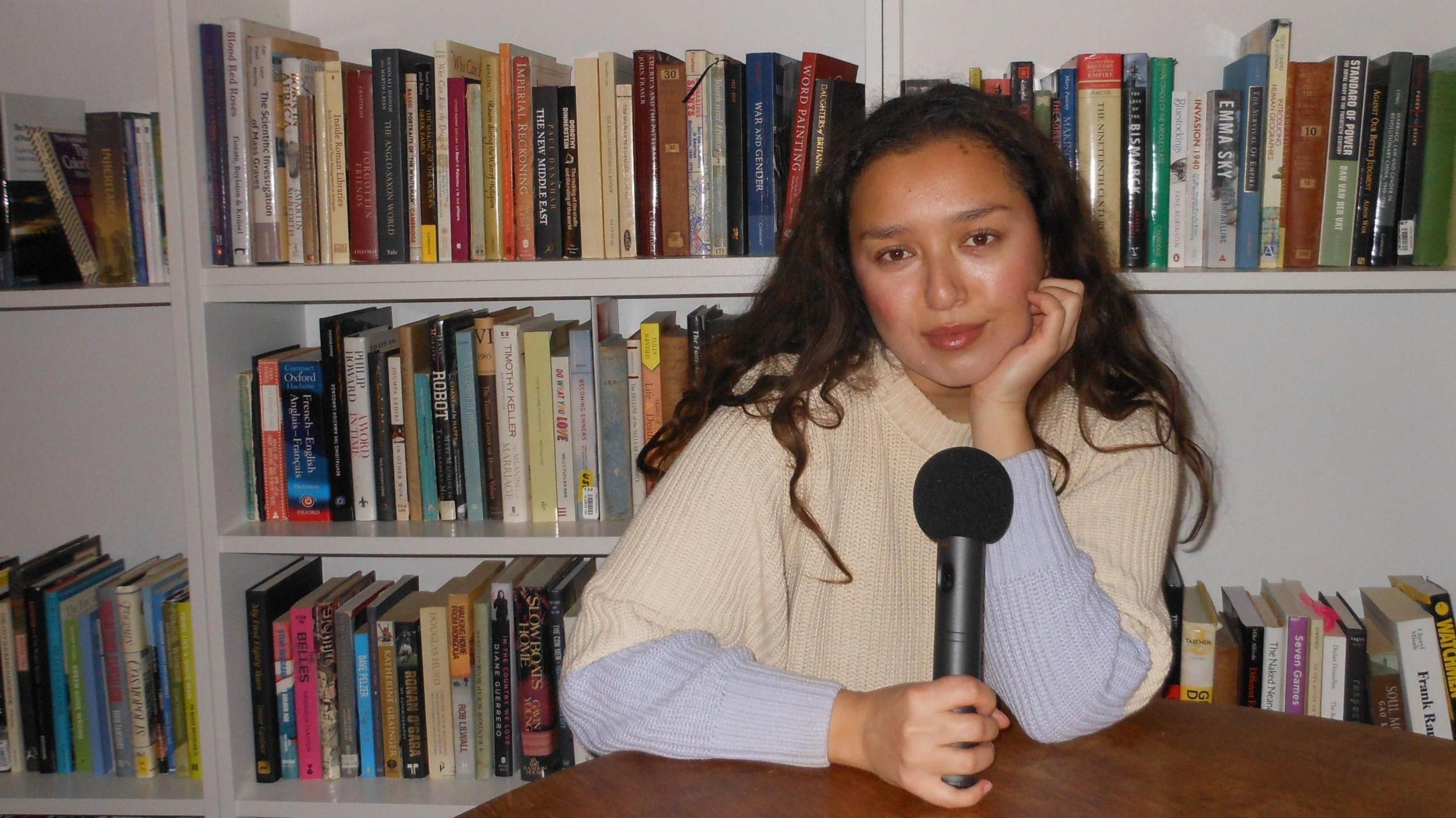 Lara Harris appearing with long dark brown hair, a woolly jumper and holding a black microphone. She is sitting at a table, with books behind her