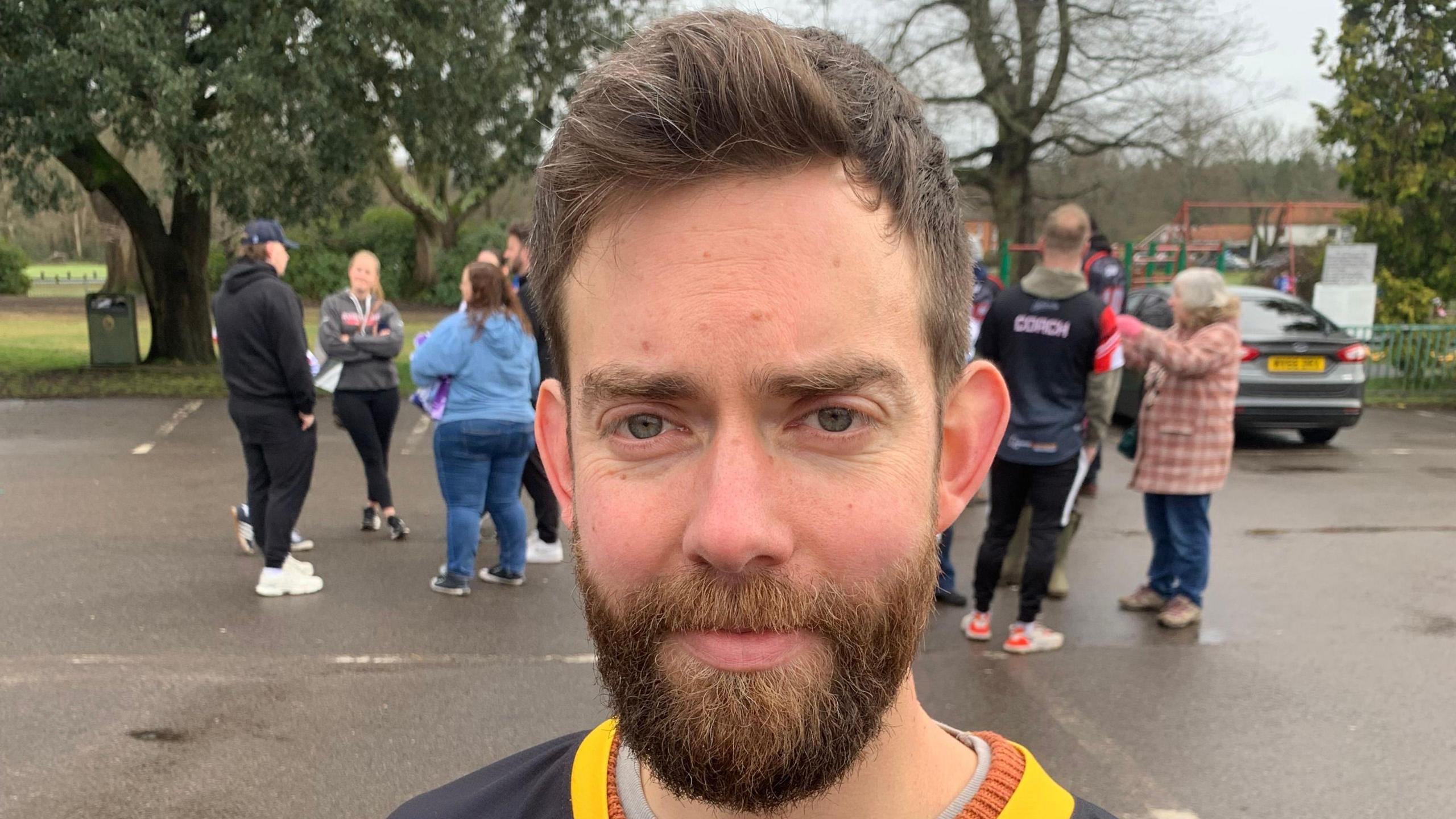 A man with a  beard looking towards the camera. A group of people are standing in the background.