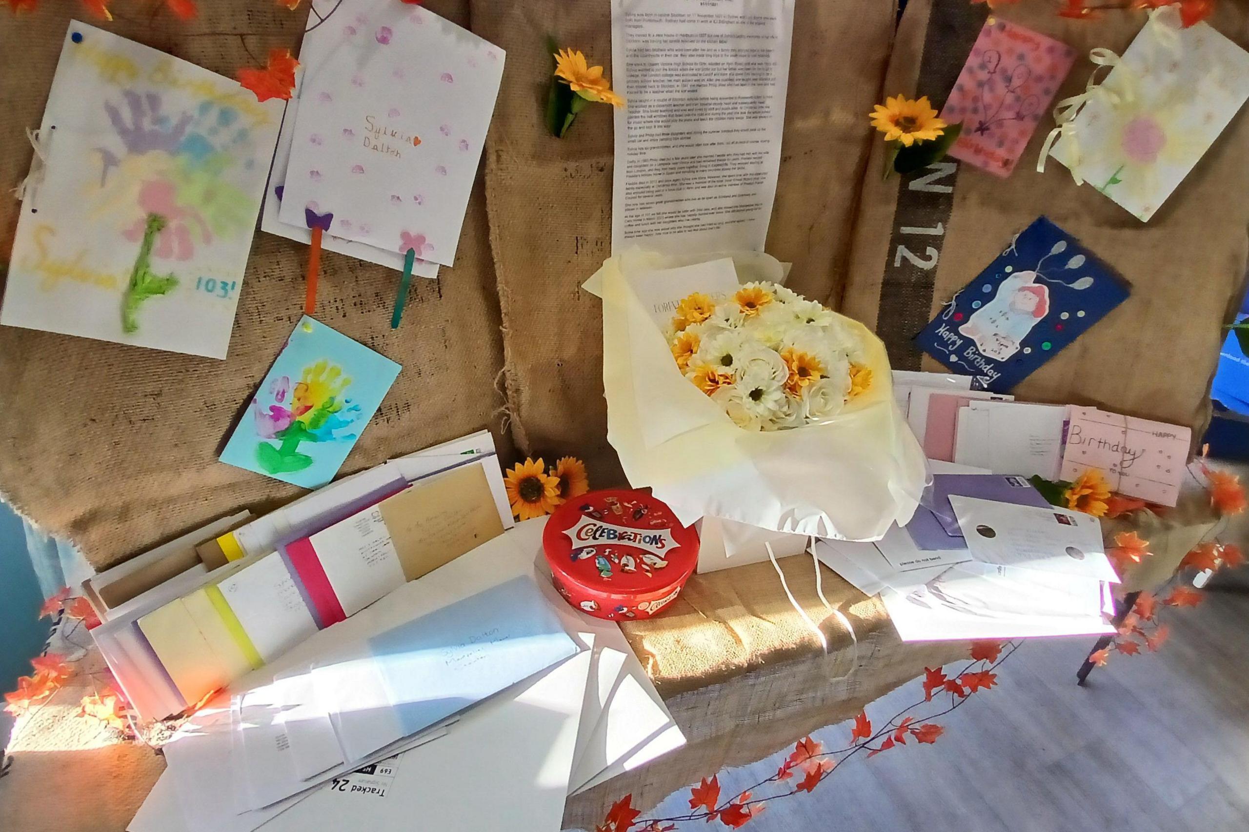 A display table decorated with brown cloth and orange leaves is covered with unopened cards and letters. There is also a bouquet of white and yellow flowers in the middle of the table, along with a tub of chocolates.