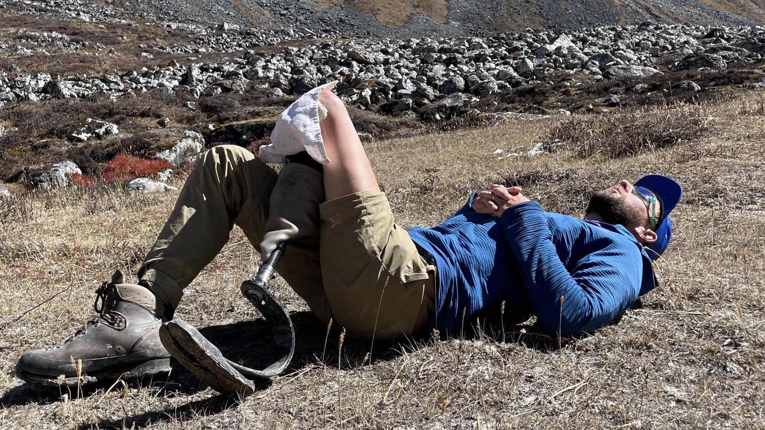 One of the men lying on his back on grass in front of a rocky area. He is wearing a blue cap and sunglasses and has his hands clasped together on his stomach,
On his right leg is a long light-brown trouser and a black boot. The lower half of his left leg is artificial. He wears a light brown short on his left left. His bare left thigh is revealed and he has a piece of white cloth on at the point where his artificial leg is fitting to his thigh. 