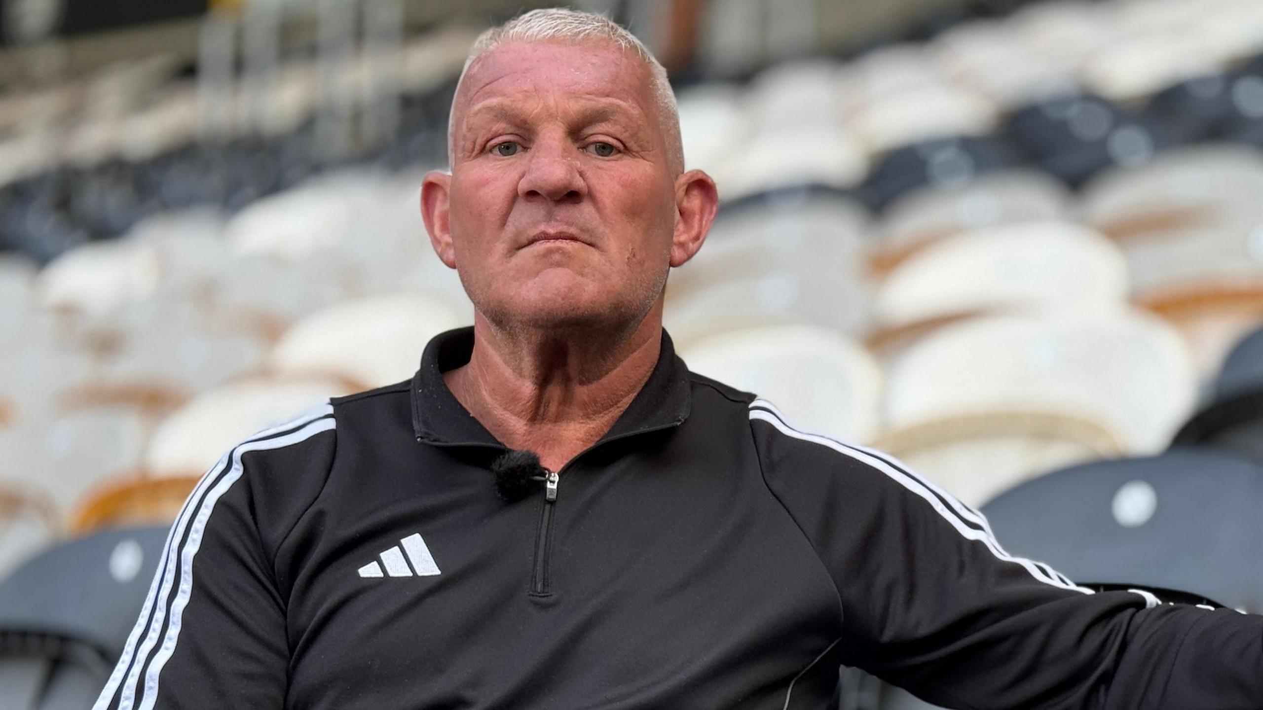 Windass, with short grey hair, is sitting on a black seat in Hull City's football stadium. He is looking directly into the camera lens and wearing a black sports top with white stripes down the side.