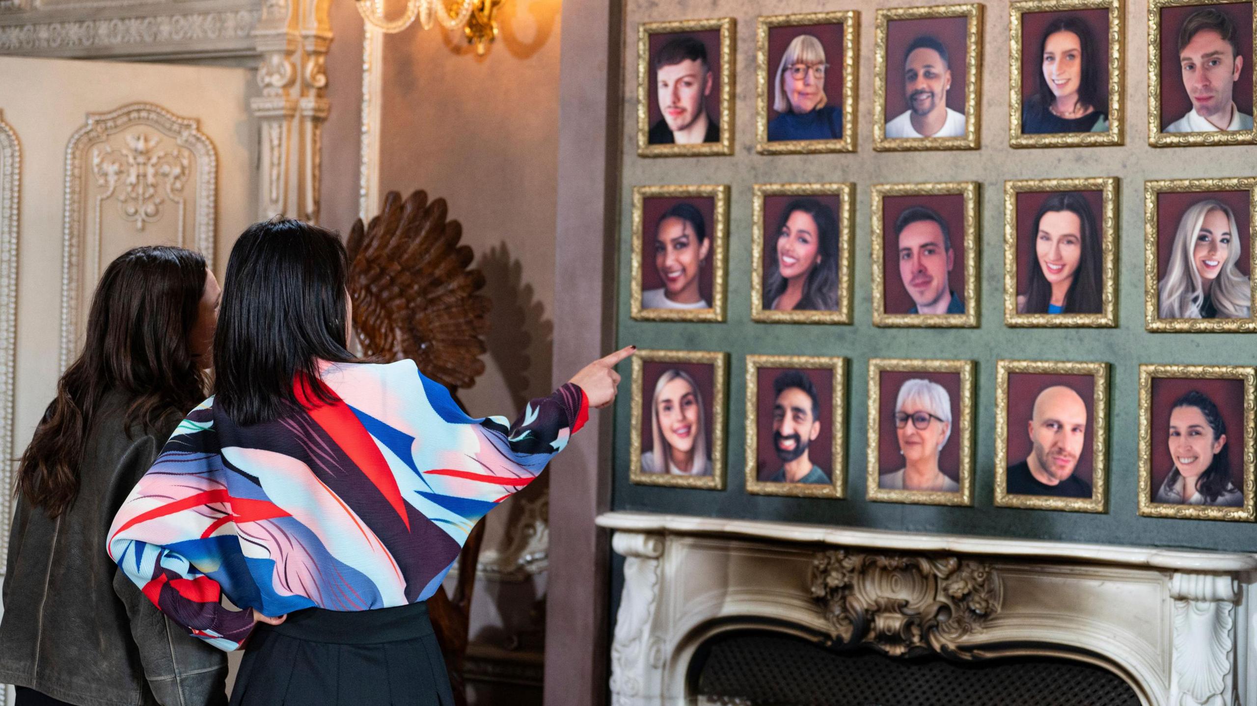 Contestants Elen and Yin look at all the portraits of the players over a mantelpiece in the Traitors castle. Each player has a small portrait on a red background in a gold frame in rows of five. 