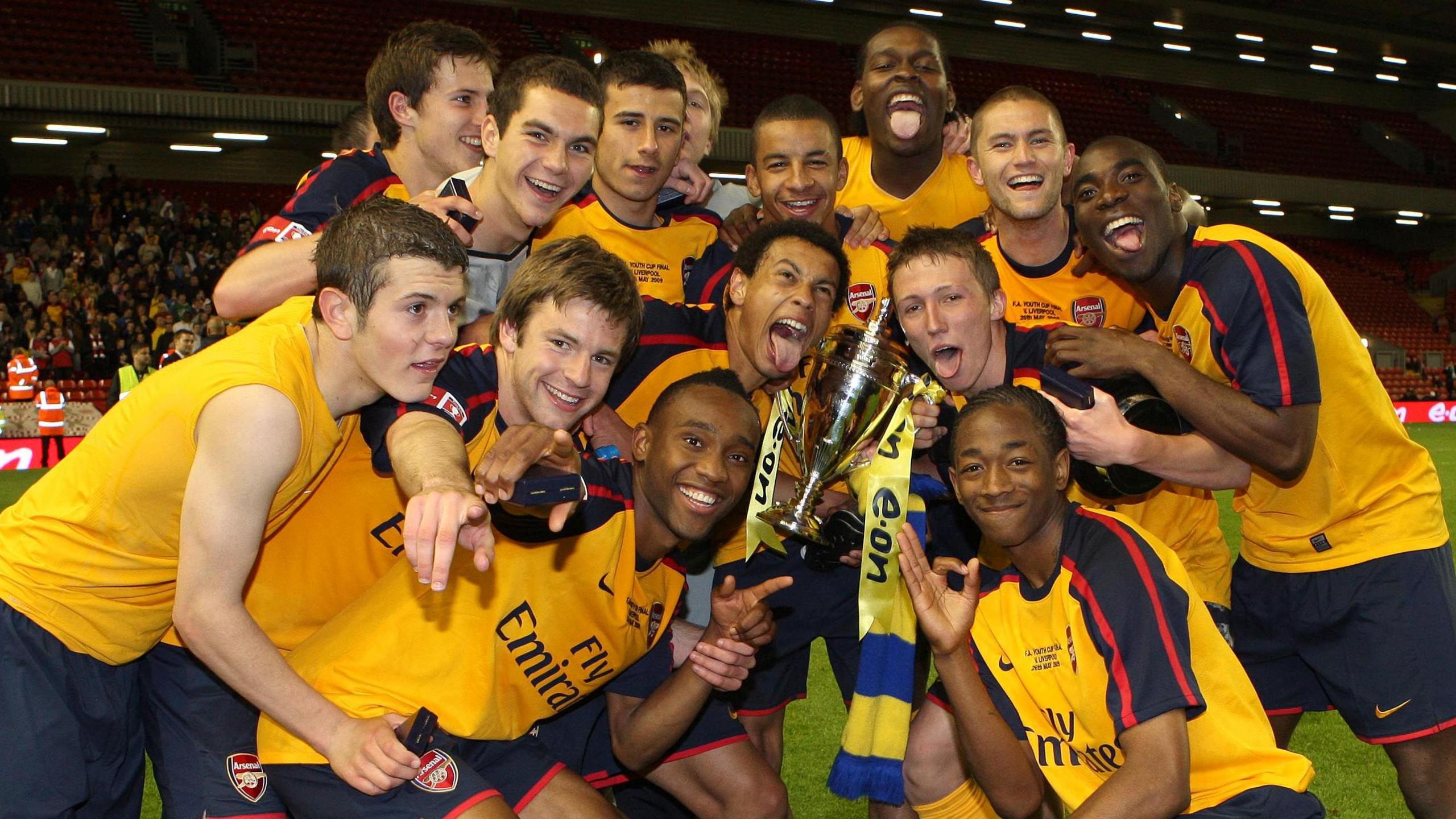 The Arsenal youth team celebrate with the FA Youth Cup trophy on the pitch. They are smiling, cheering and poking their tongues out while wearing a yellow jersey with dark blue shorts.