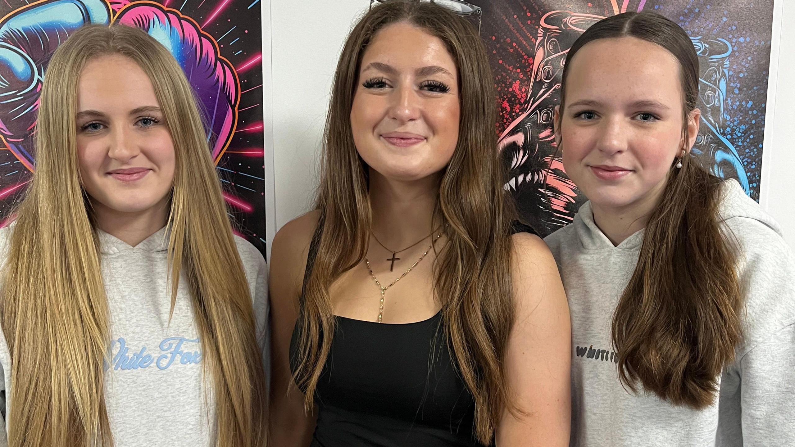 The three sisters standing side-by-side smiling at camera. Two of them are wearing grey tops and one in the middle is wearing a black top. The sister on the right has her hair tied and the others have their hair out.