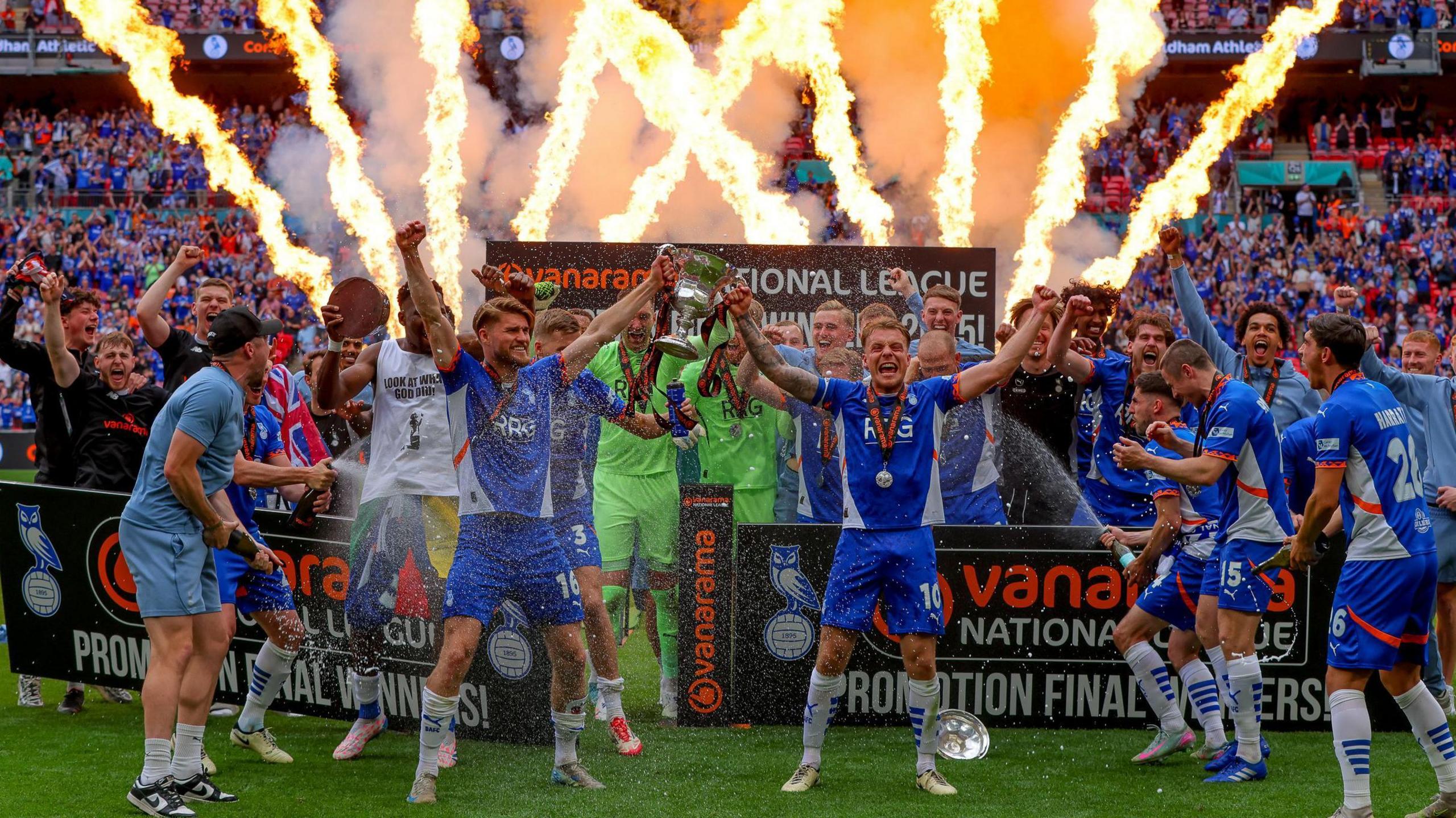 Oldham Athletic players celebrate winning the National League play-off final and promotion to the English Football League on the pitch at Wembley with fireworks streaking into the sky behind them.