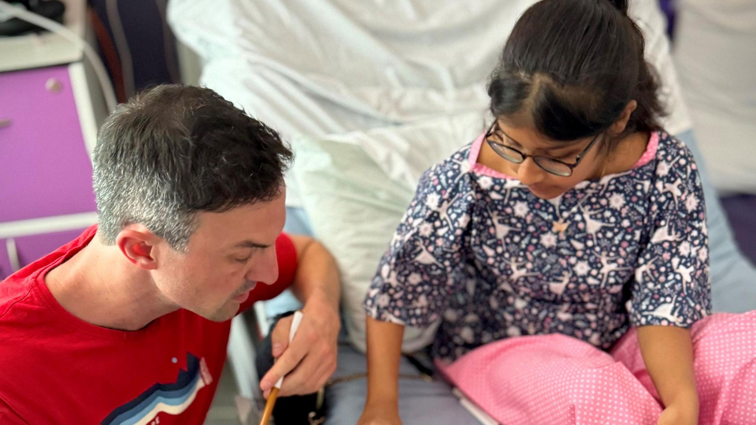Jamie kneeling beside the bed of a girl who is in hospital. They are drawing together. 