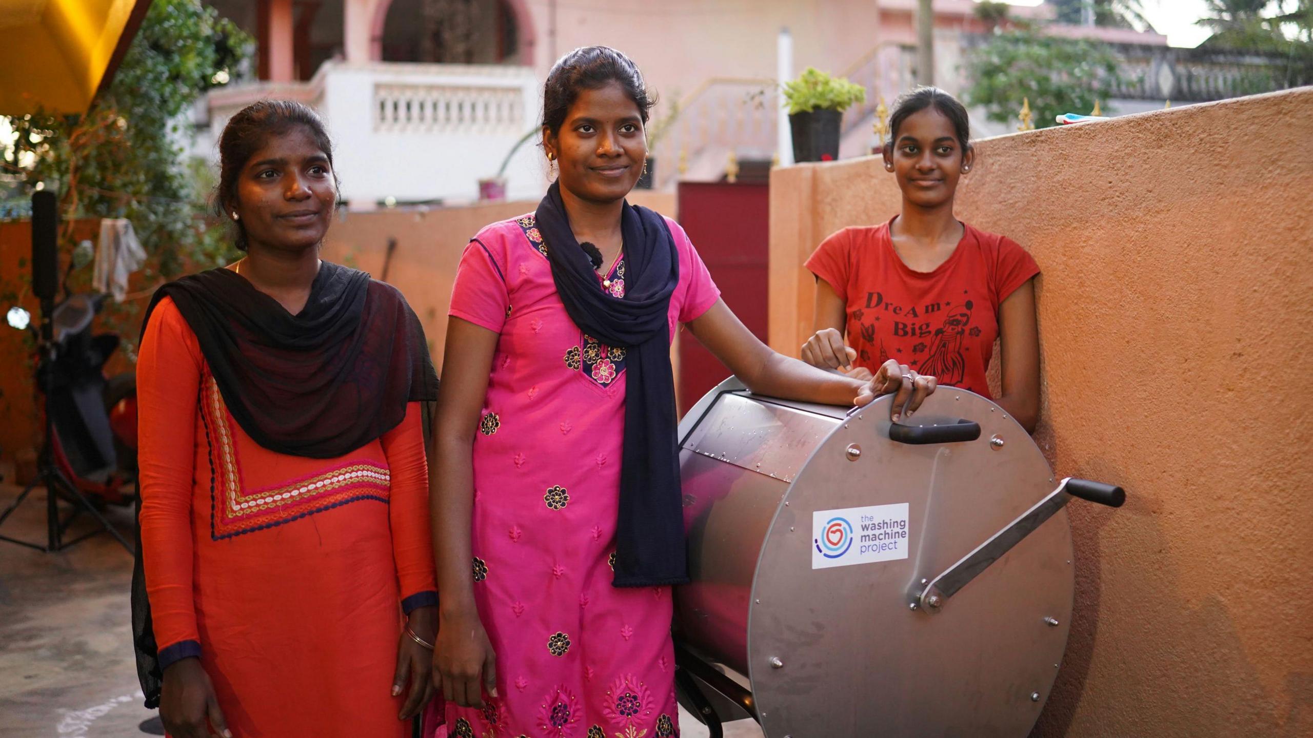 Three women pictured smiling in a courtyard area with a Divya washing machine.  