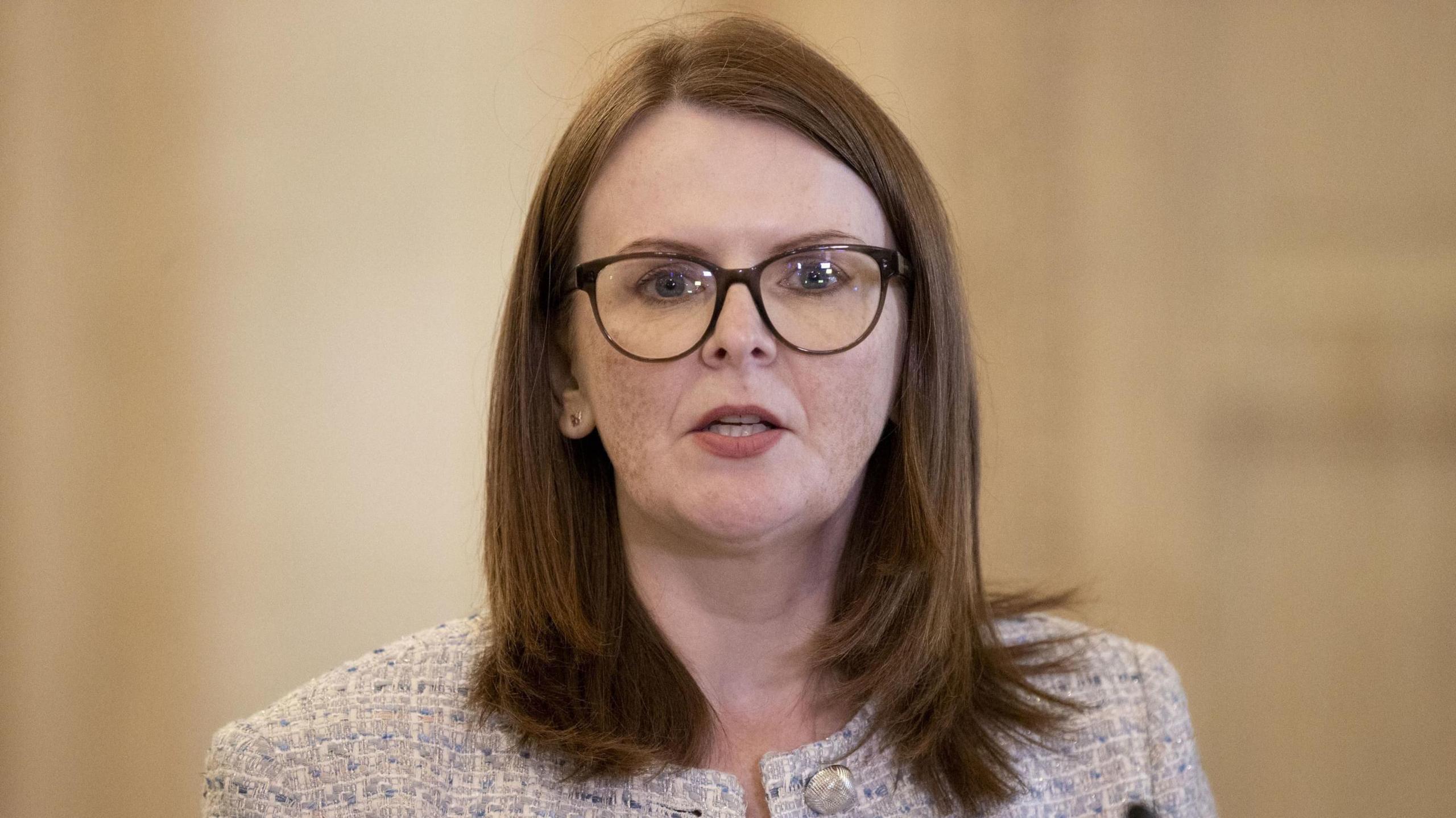 Caoimhe Archibald wearing glasses and a cream tweed jacket.  She is looking to camera and is mid-speech