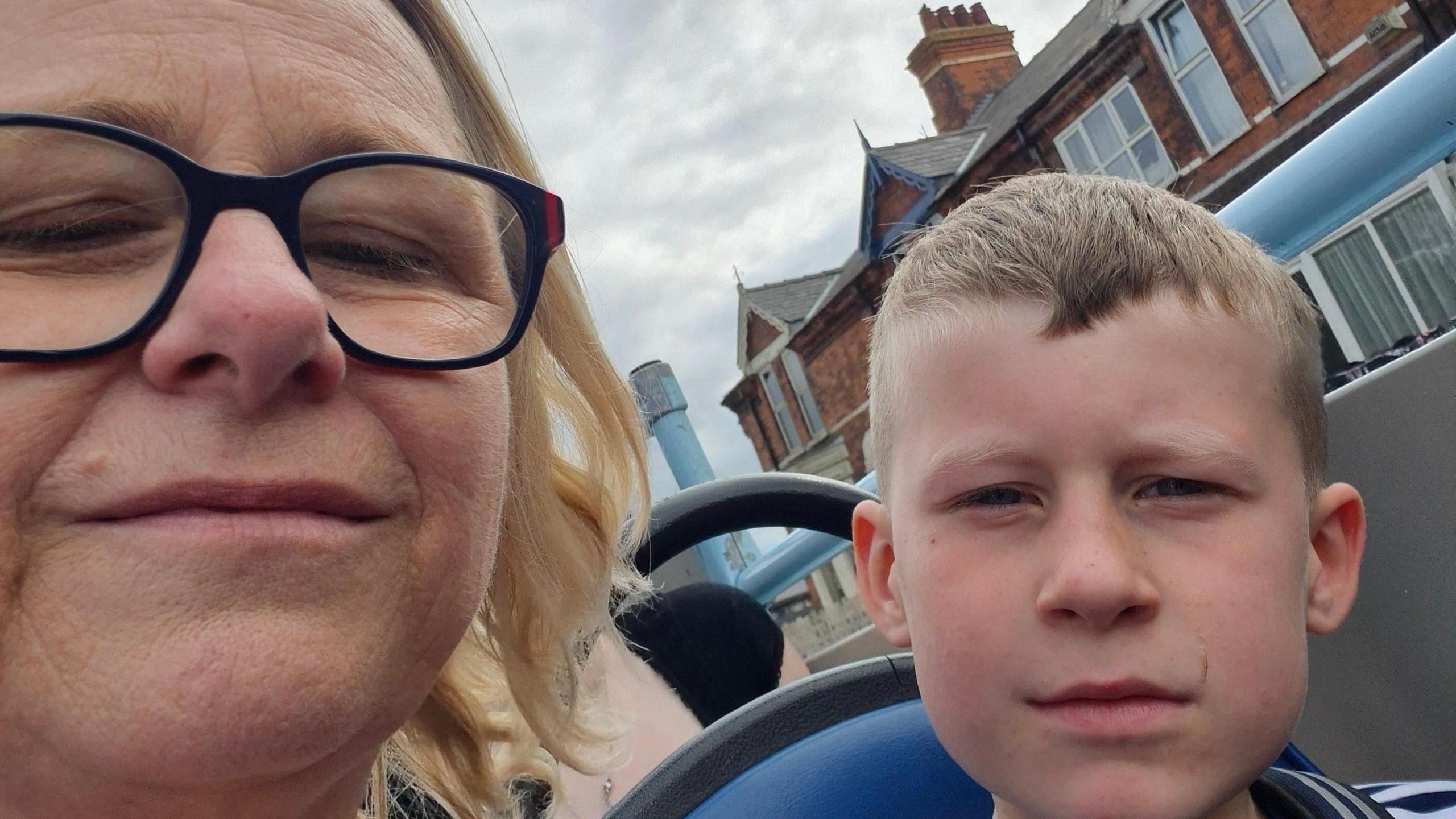 A head and shoulders selfie-shot of a blonde woman with short hair and glasses on an open top bus with her seven year old grandson who is sat next to her. 