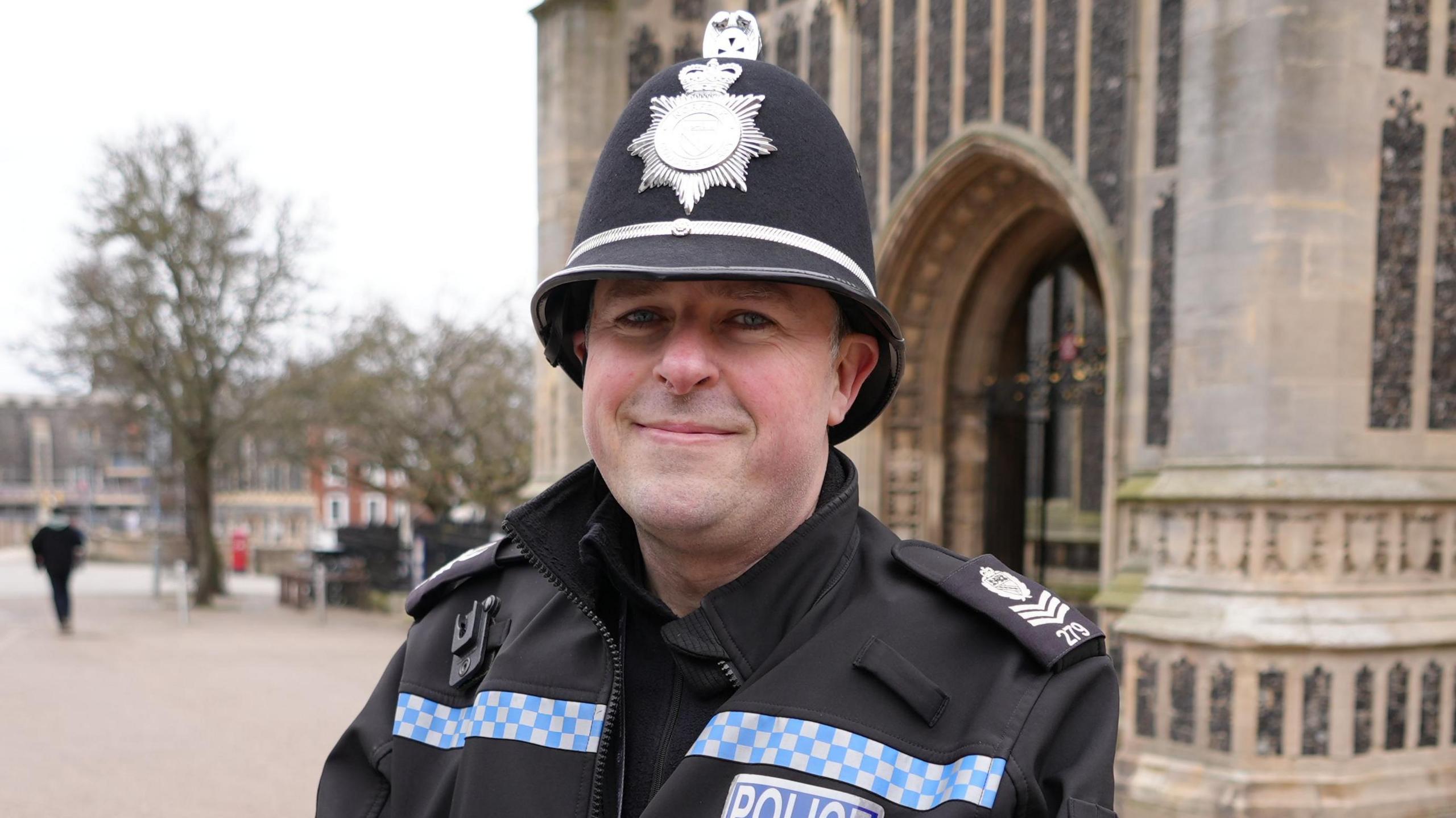 Sgt Chris Clay. A man who is looking at the camera and smiling. He is a police officer who is dressed in a black police uniform with a coat. He is also wearing a police hat and is standing outside a church. 