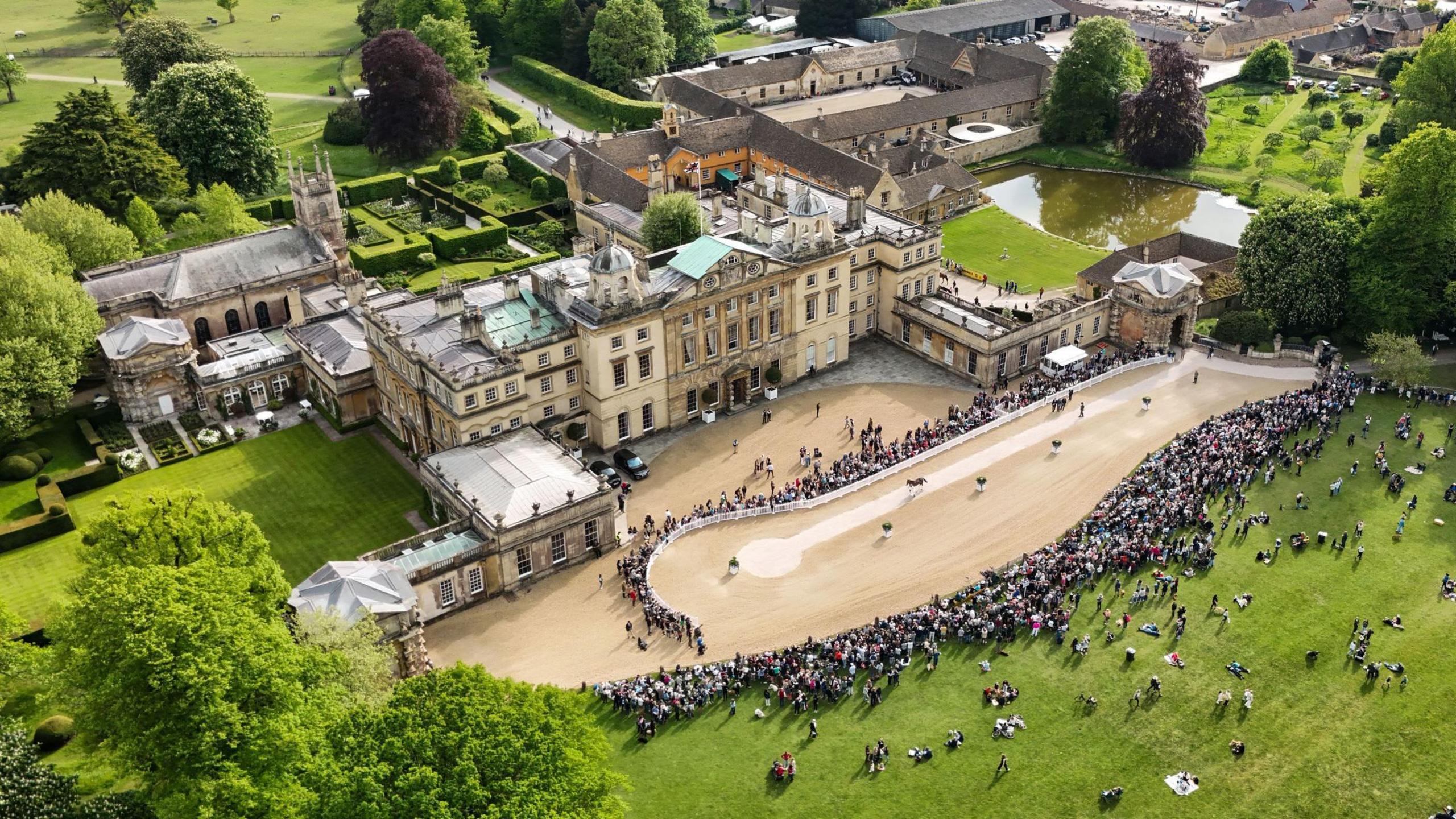 An aerial view of the Badminton Horse trials taken from hundreds of feet in the air shows the main house and the parade ring in front of it with hundreds of spectators looking on