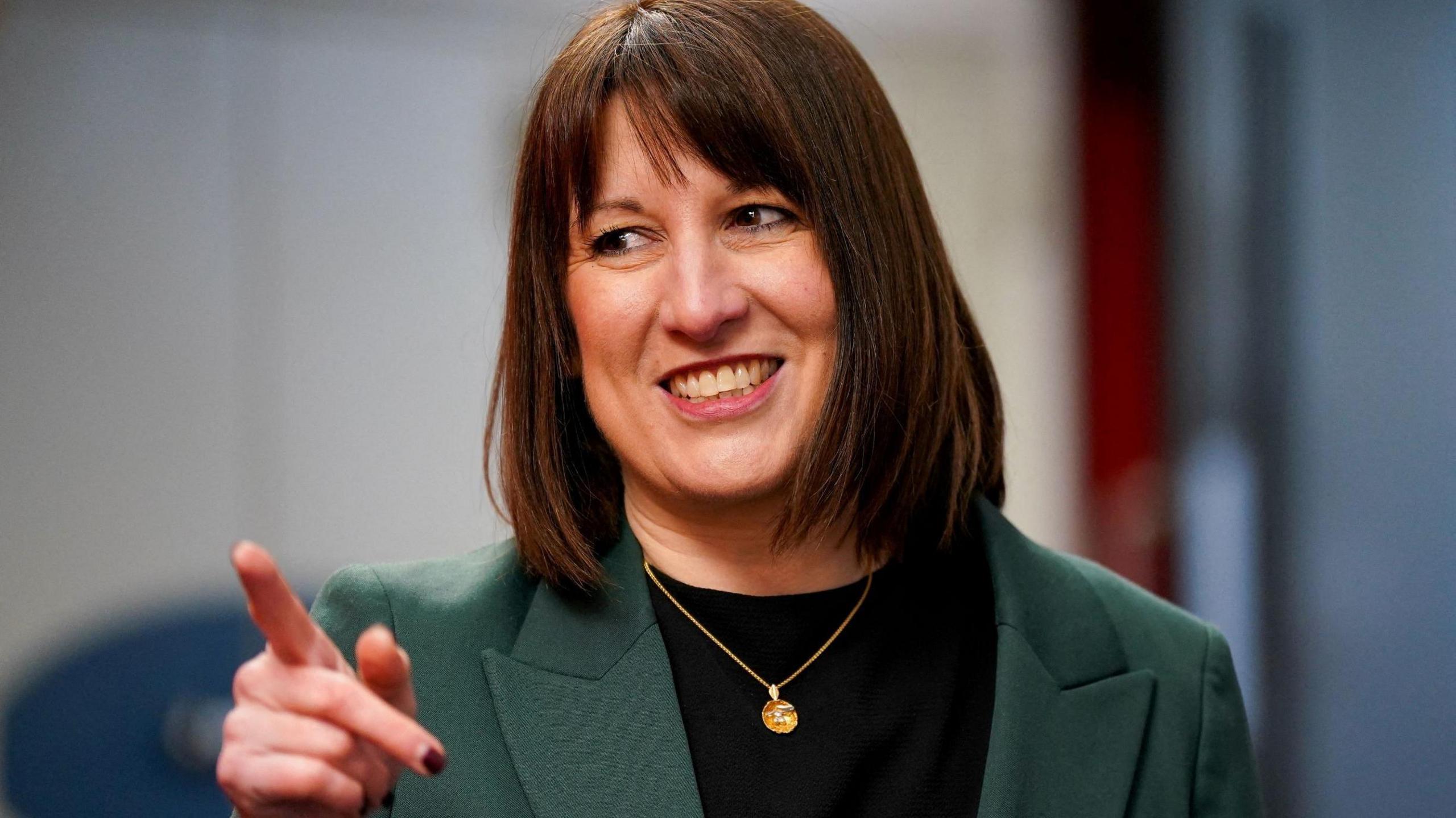 Rachel Reeves, with brown hair cut in a bob, points and smiles. She is photographed from the chest up and wearing a black top and green jacket. She is also wearing a gold pendant.