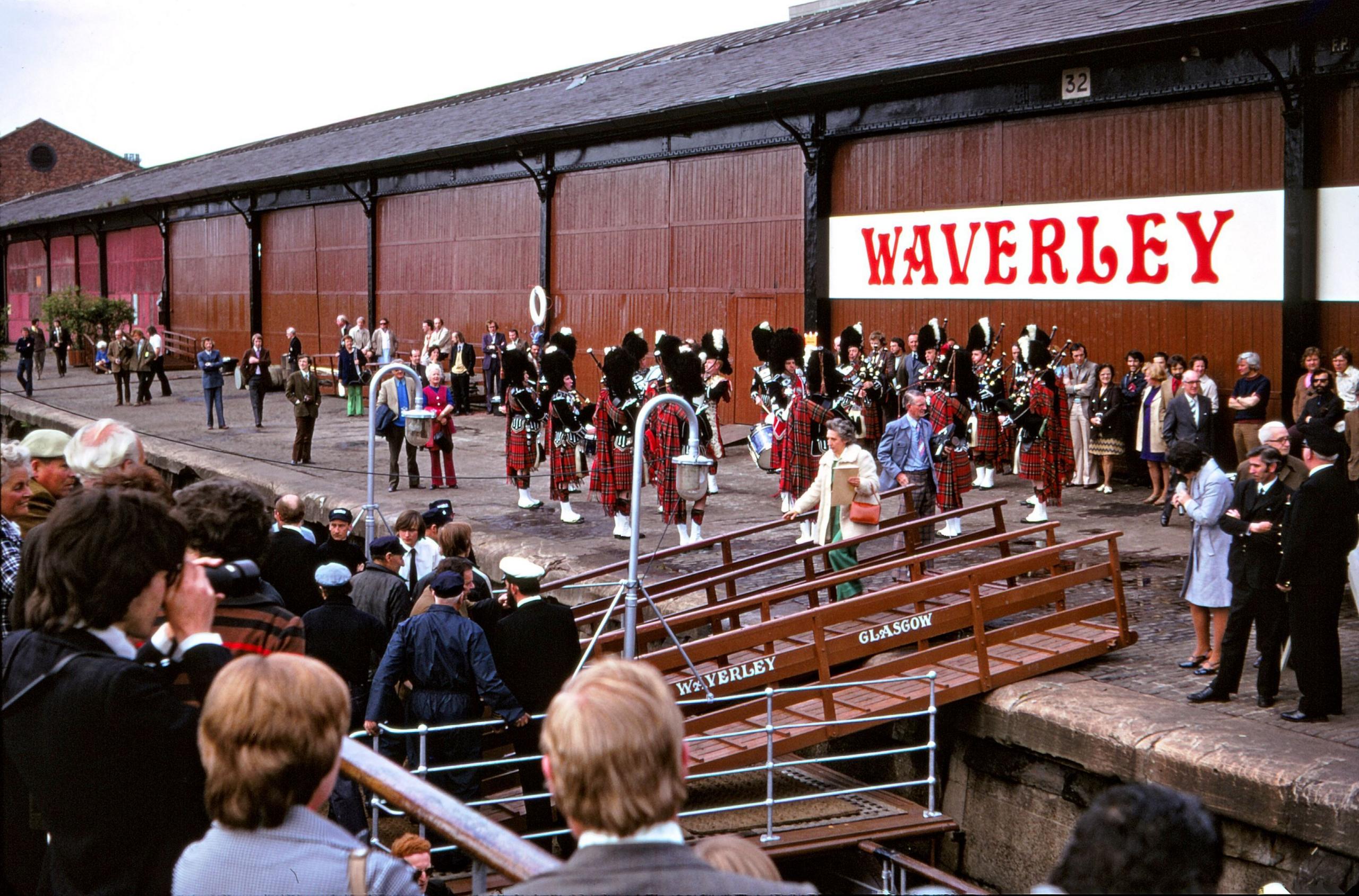 A crown gathers on the Waverley as we look across to the quayside where a pipe band is playing to see it off. A well-dressed woman is walking across the passerelle and onto the boat.