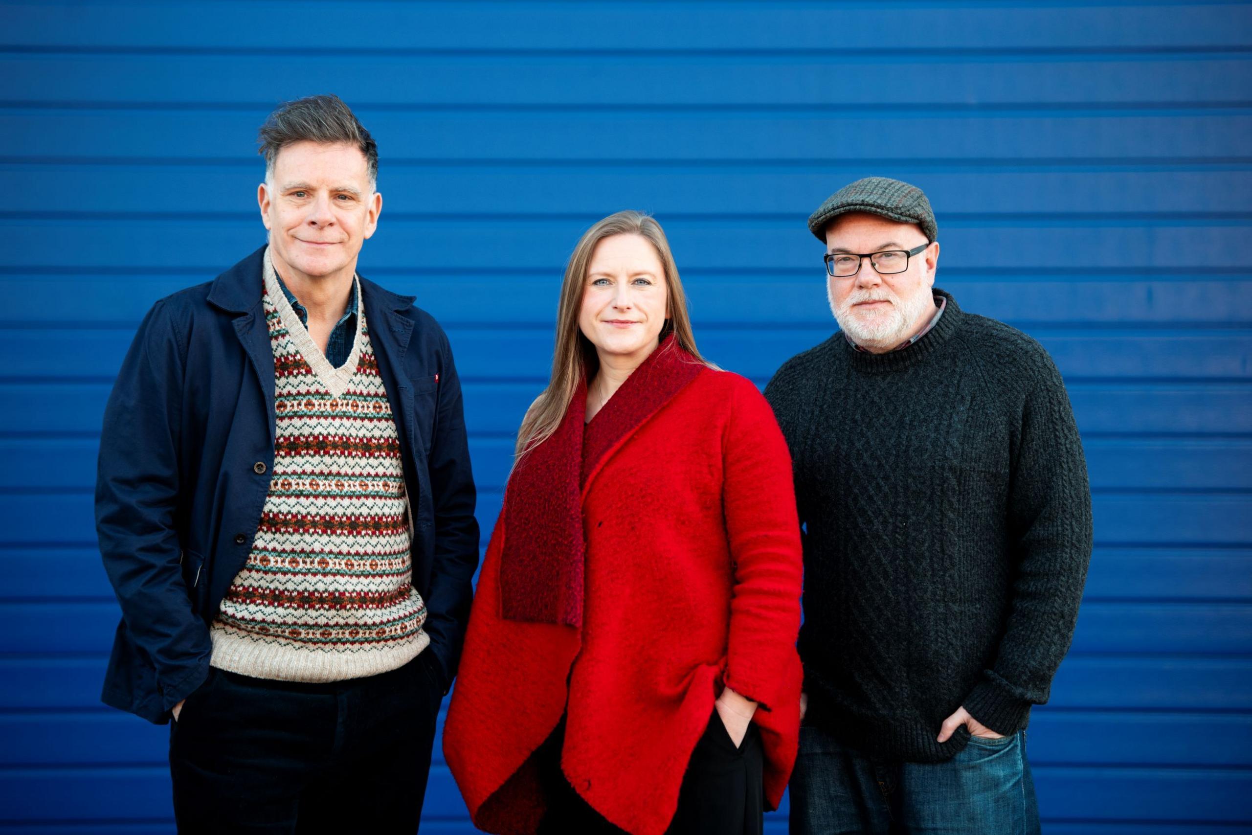 Ricky Ross, left, wearing a multi-coloured top under a dark blue jacket. Frances Poet, centre, wearing a red top. Dominic Hill, right, wearing a multi-coloured dark flat cap, glasses and a dark jumper. All three are standing in front of a blue background.