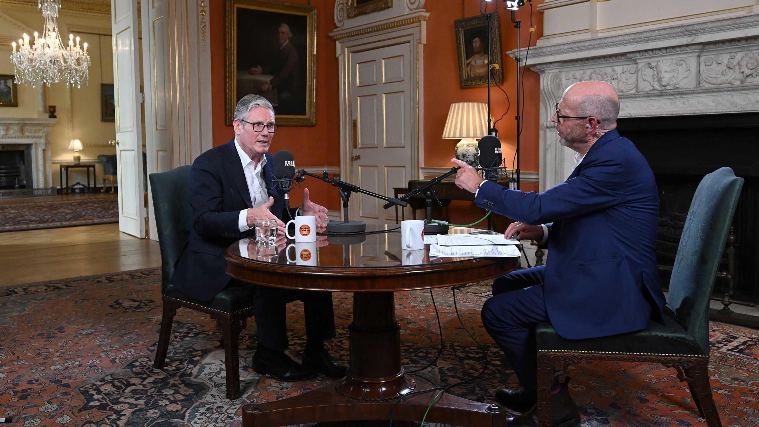 Two men (Sir Keir Starmer and Nick Robinson) sit opposite each other at a table with microphones on top positioned towards them